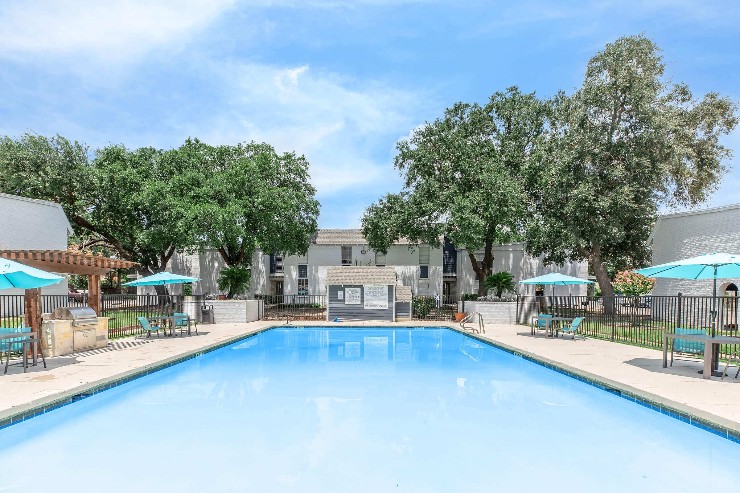 A clear blue swimming pool surrounded by a lounge area with green umbrellas. In the background, there are large trees and white apartment buildings, creating a serene outdoor space. The sky is bright with a few clouds, enhancing the inviting atmosphere of the pool area.