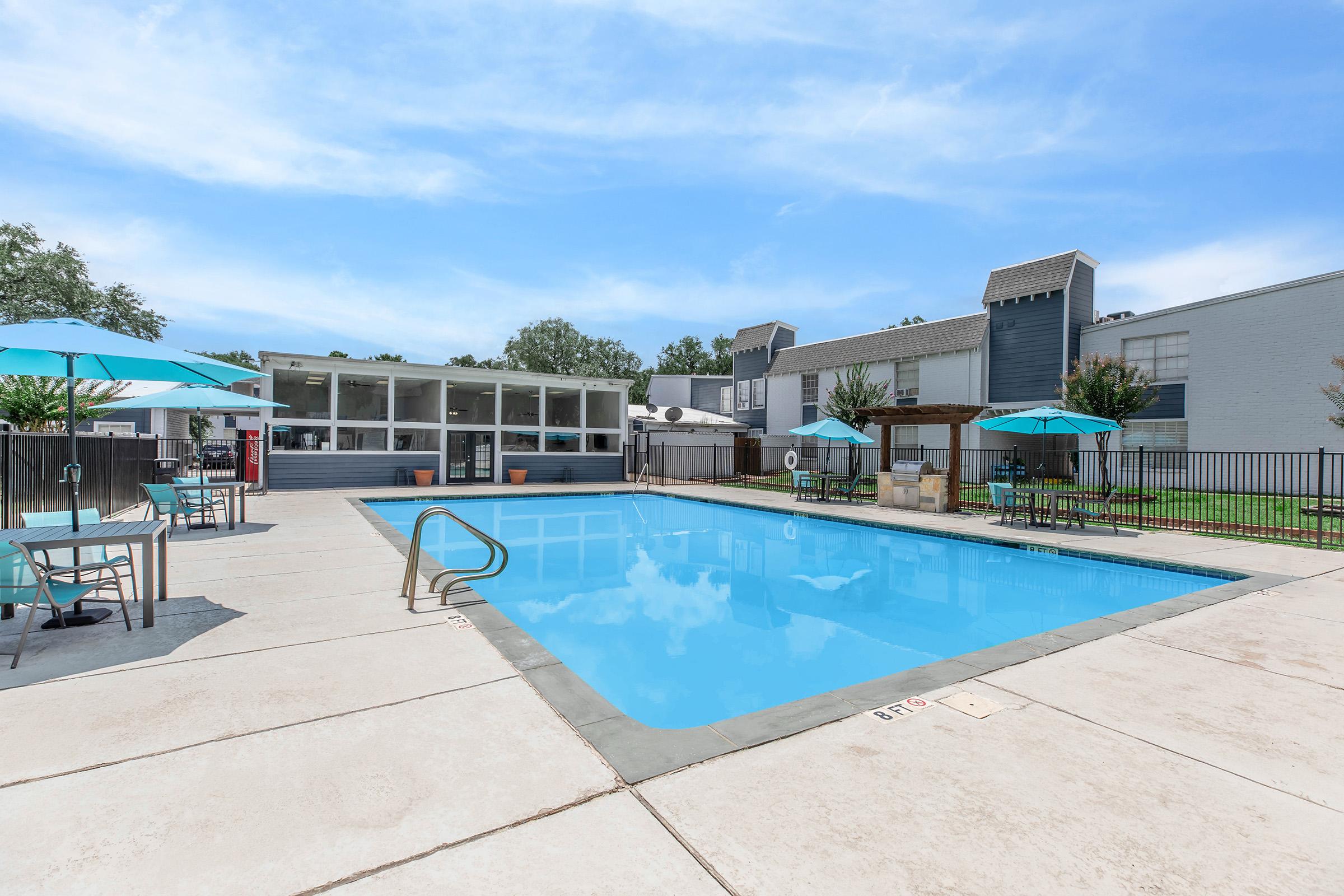 A clean and inviting swimming pool surrounded by lounge chairs and tables with teal umbrellas. The area features a light-colored concrete deck with a chain-link fence. In the background, there is a building with large windows and additional outdoor seating, all set against a clear blue sky.