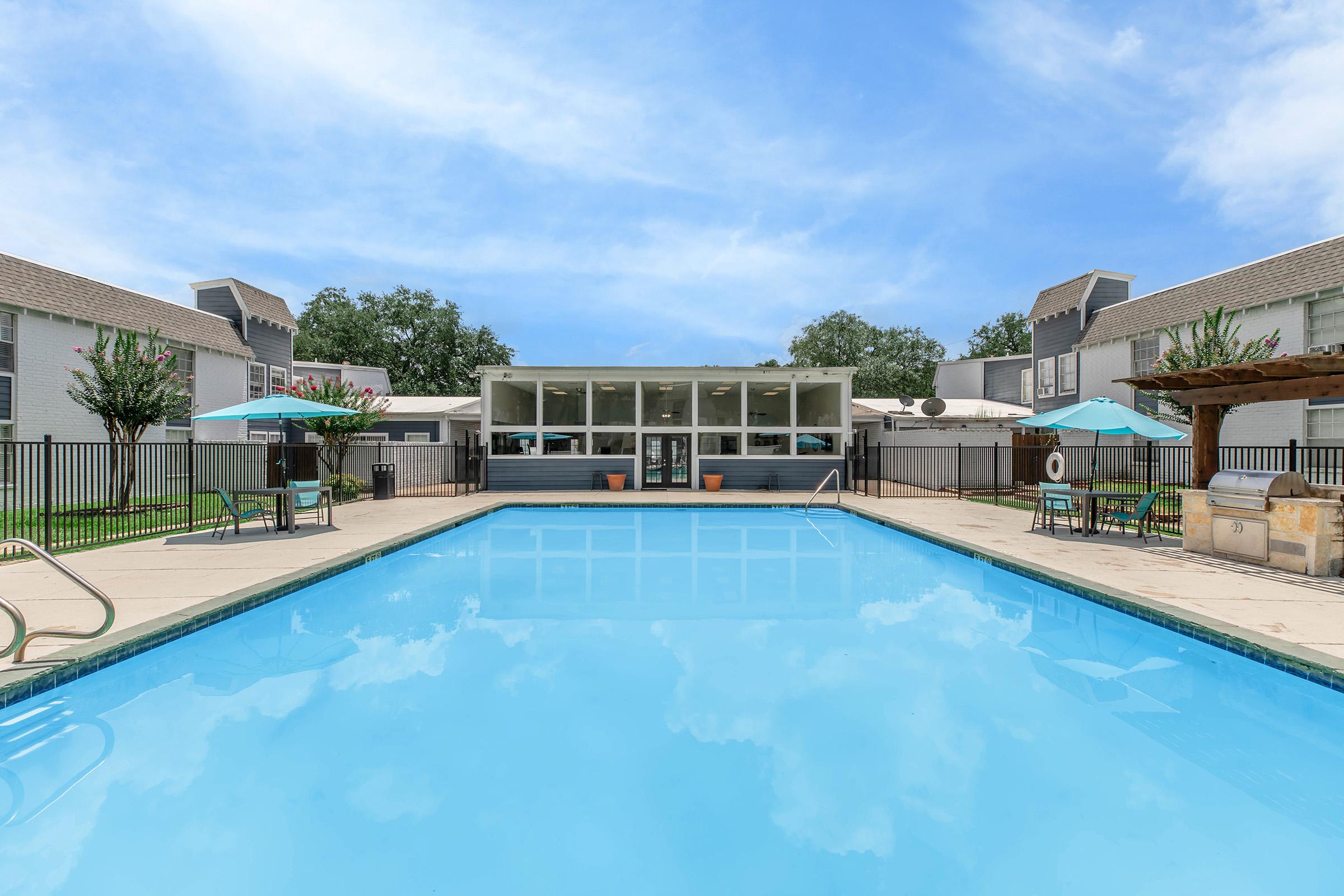 Swimming pool with clear blue water surrounded by lounge chairs and umbrellas. A modern pool house with large windows is visible in the background, alongside greenery and well-maintained landscaping. The sky is bright with few clouds.