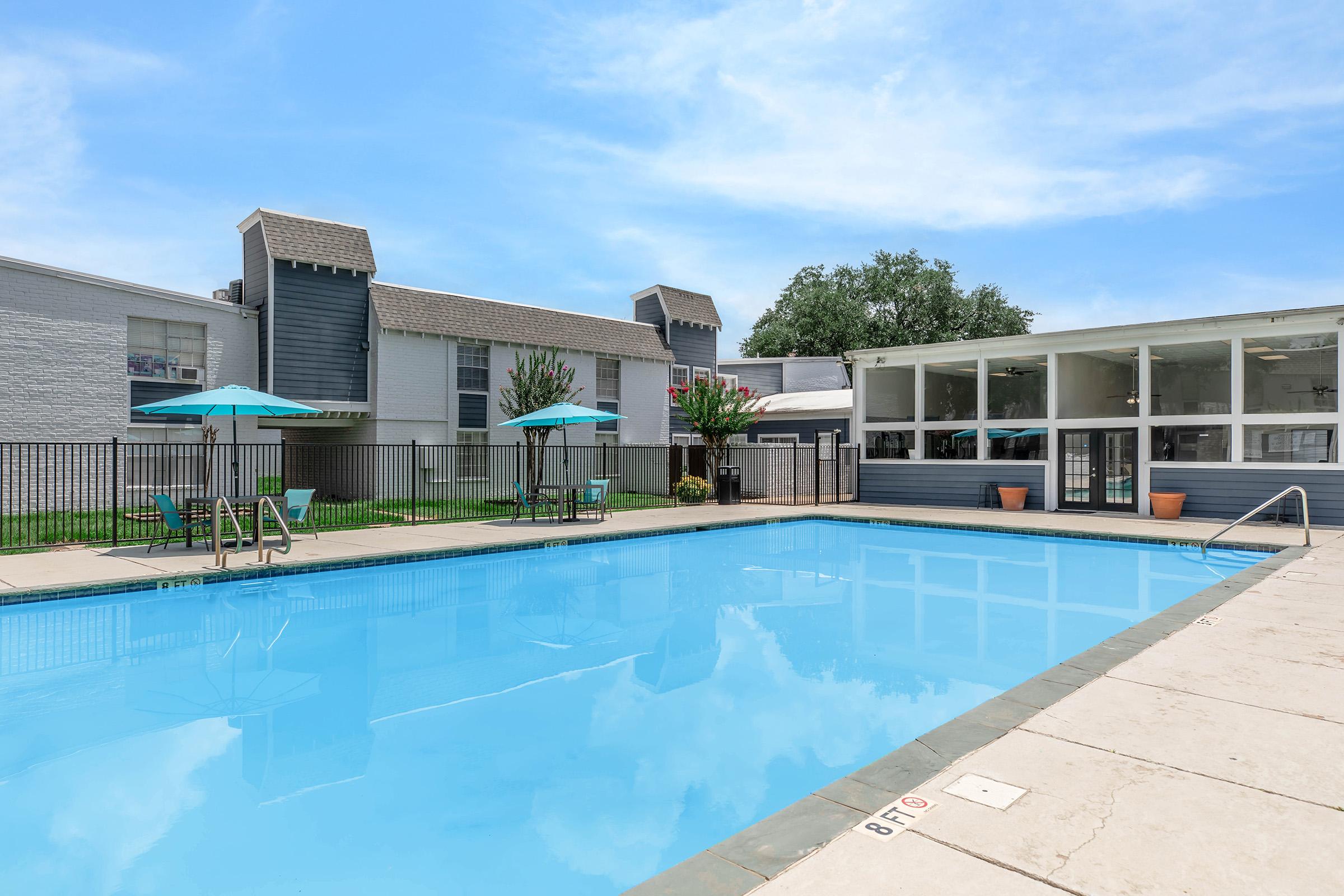 A blue swimming pool surrounded by a concrete deck, with lounge chairs and teal umbrellas. In the background, there are two-story residential buildings and some greenery. The sky is clear with a few clouds, creating a bright and inviting atmosphere.