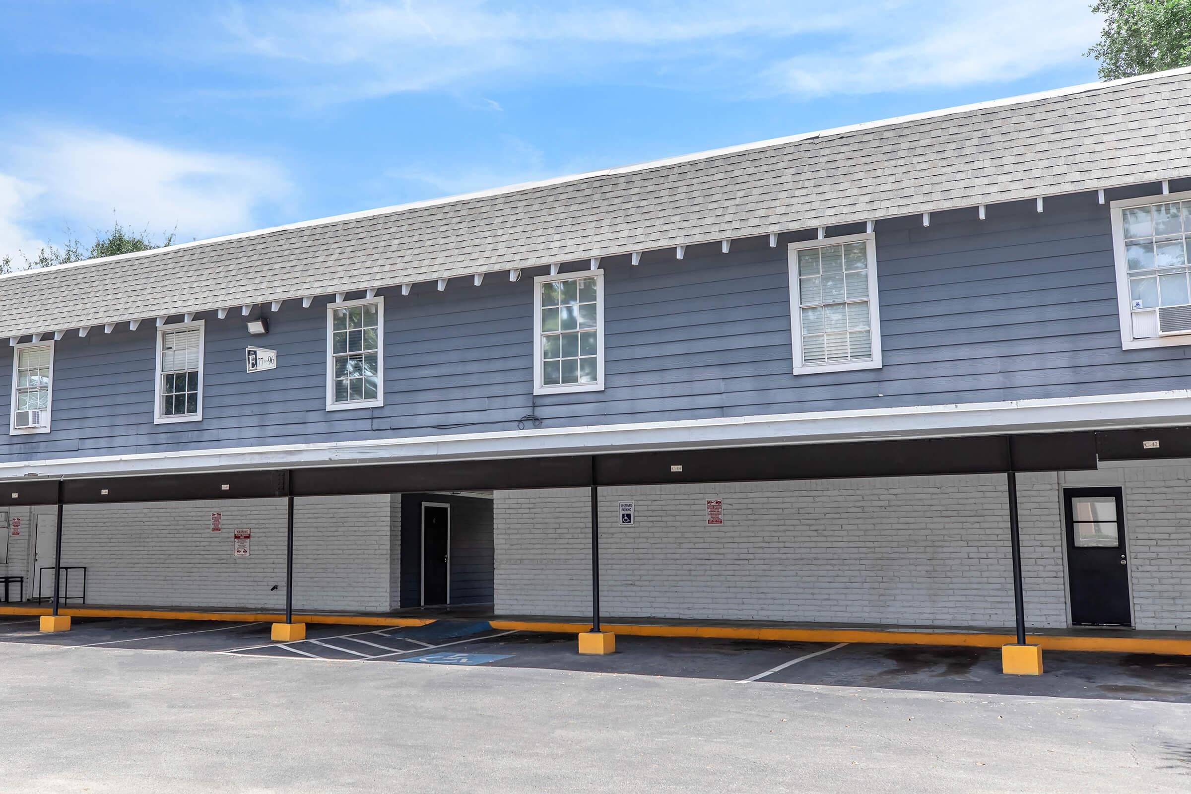 Two-story motel building with a gray exterior. The upper level has several windows, while the ground level features a wide porch area. There are parking spaces in front with a few distinct markings. The sky is clear and blue, adding to the overall bright appearance of the scene.