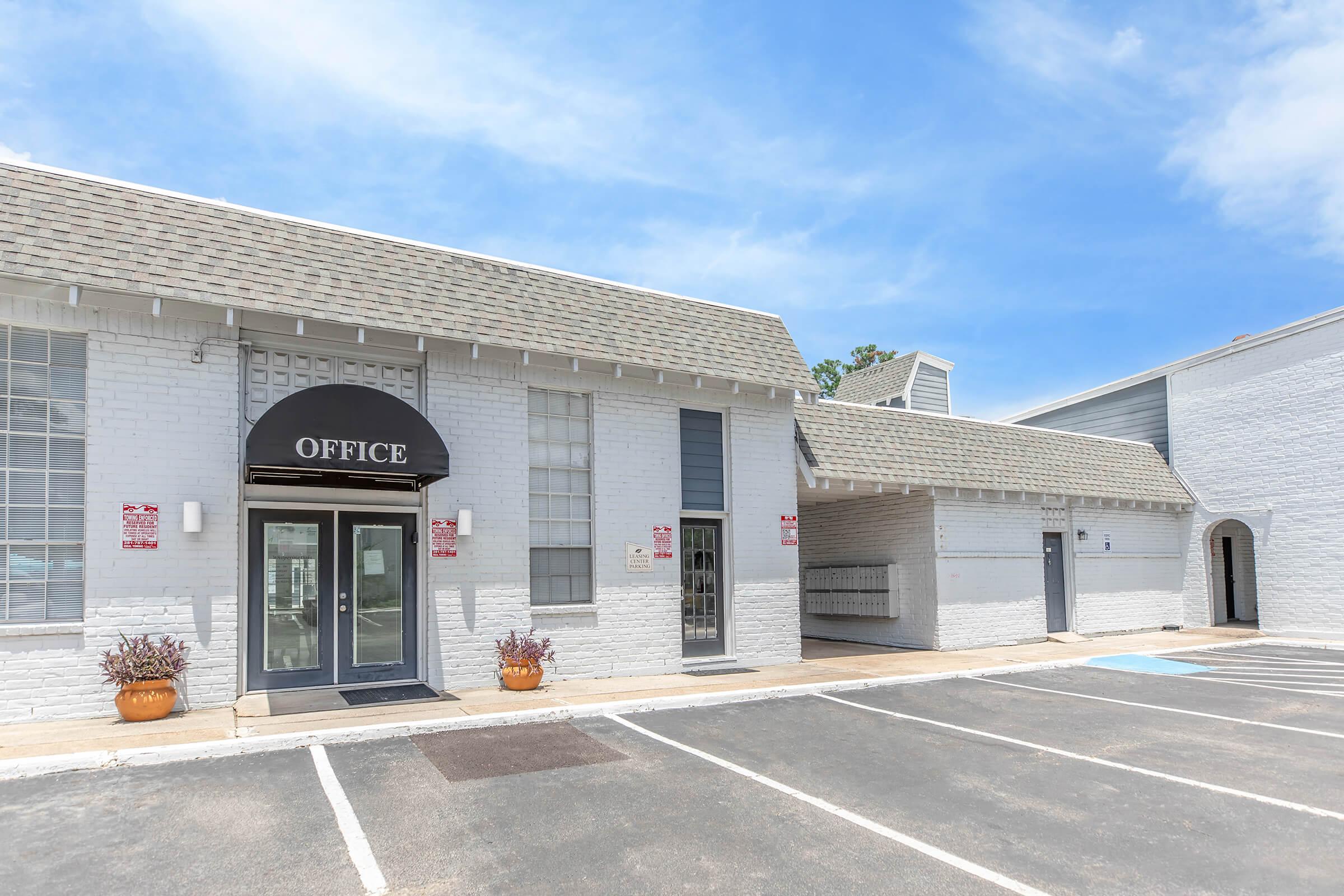 Exterior view of a white brick building featuring an office entrance with a black awning labeled "OFFICE." The surrounding area includes a parking lot and blue sky. There are potted plants near the entrance, and additional buildings can be seen in the background.