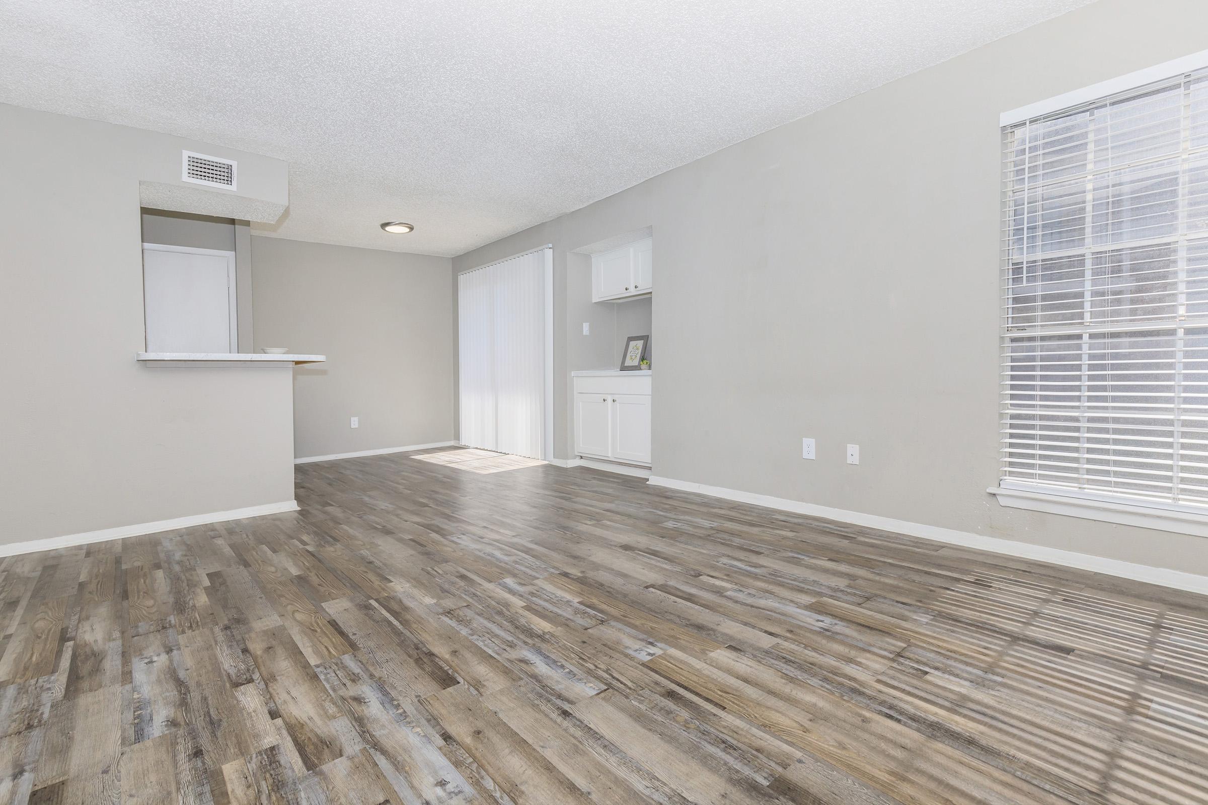 A spacious, well-lit room with light gray walls and a wood-like floor. To the left, there's an open kitchen area with a counter, and to the right, a window with blinds allowing natural light. The space is uncluttered, creating a sense of airiness and convenience for various uses.