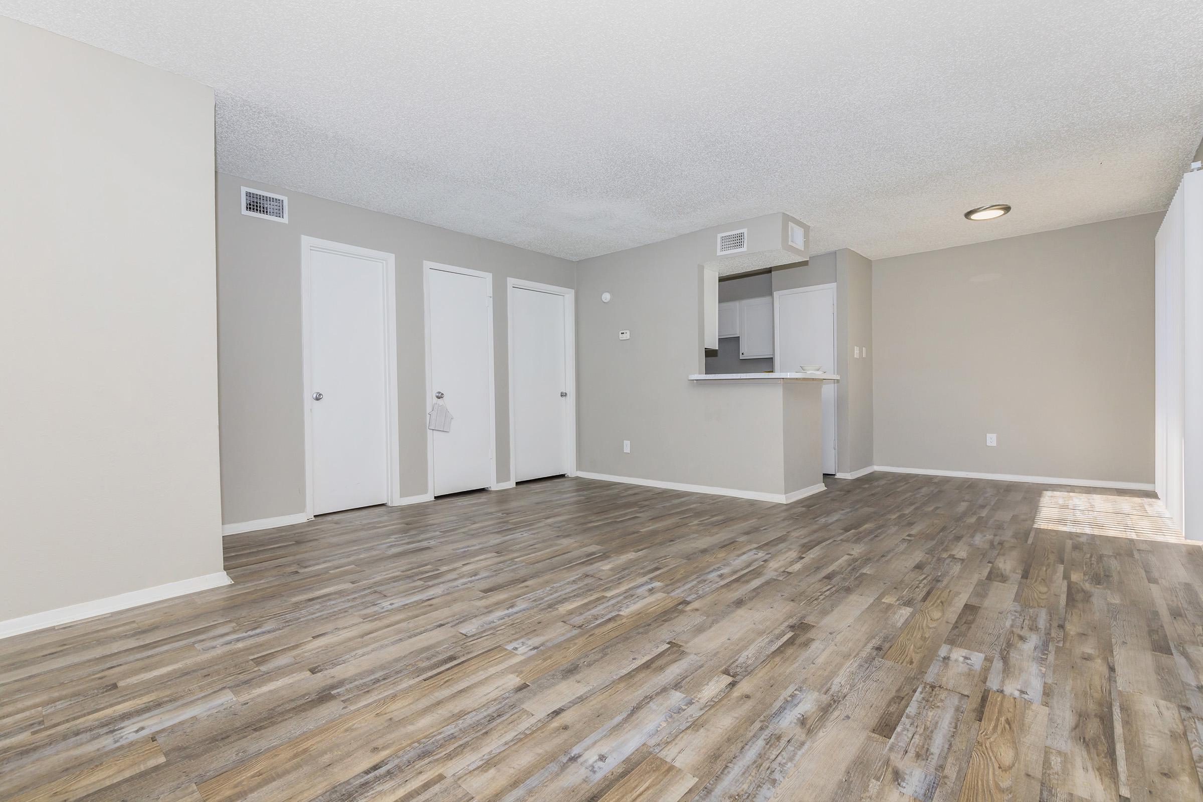 Spacious, bright living area with light wood-finish flooring. Gray walls create a modern look. Three closed white doors on the left, leading to additional rooms or storage. A small kitchen area is visible in the background with a bar counter, complementing the open layout. Natural light enhances the inviting atmosphere.