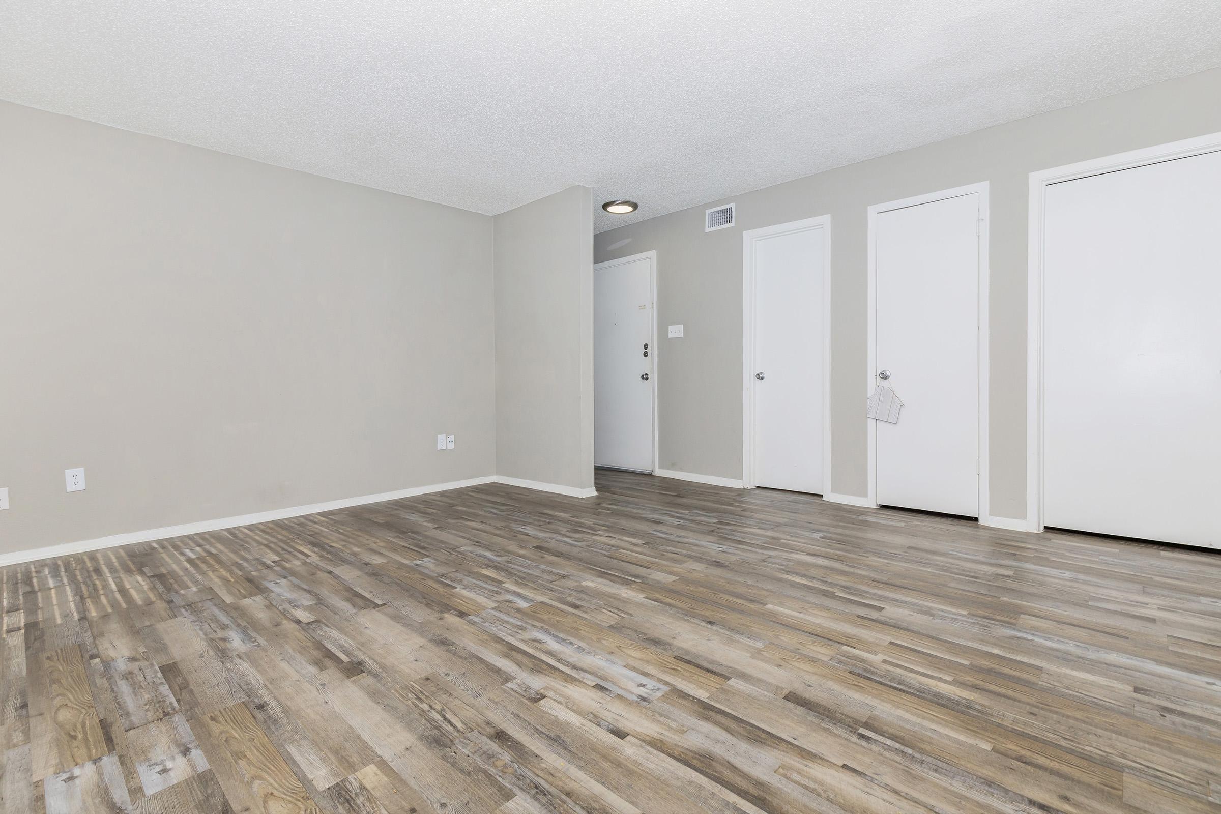 A spacious and empty room featuring light-colored walls and a textured ceiling. The floor has a wood-like finish, and there are three white doors along one wall. The entrance door is visible in the background, contributing to an open and airy atmosphere.