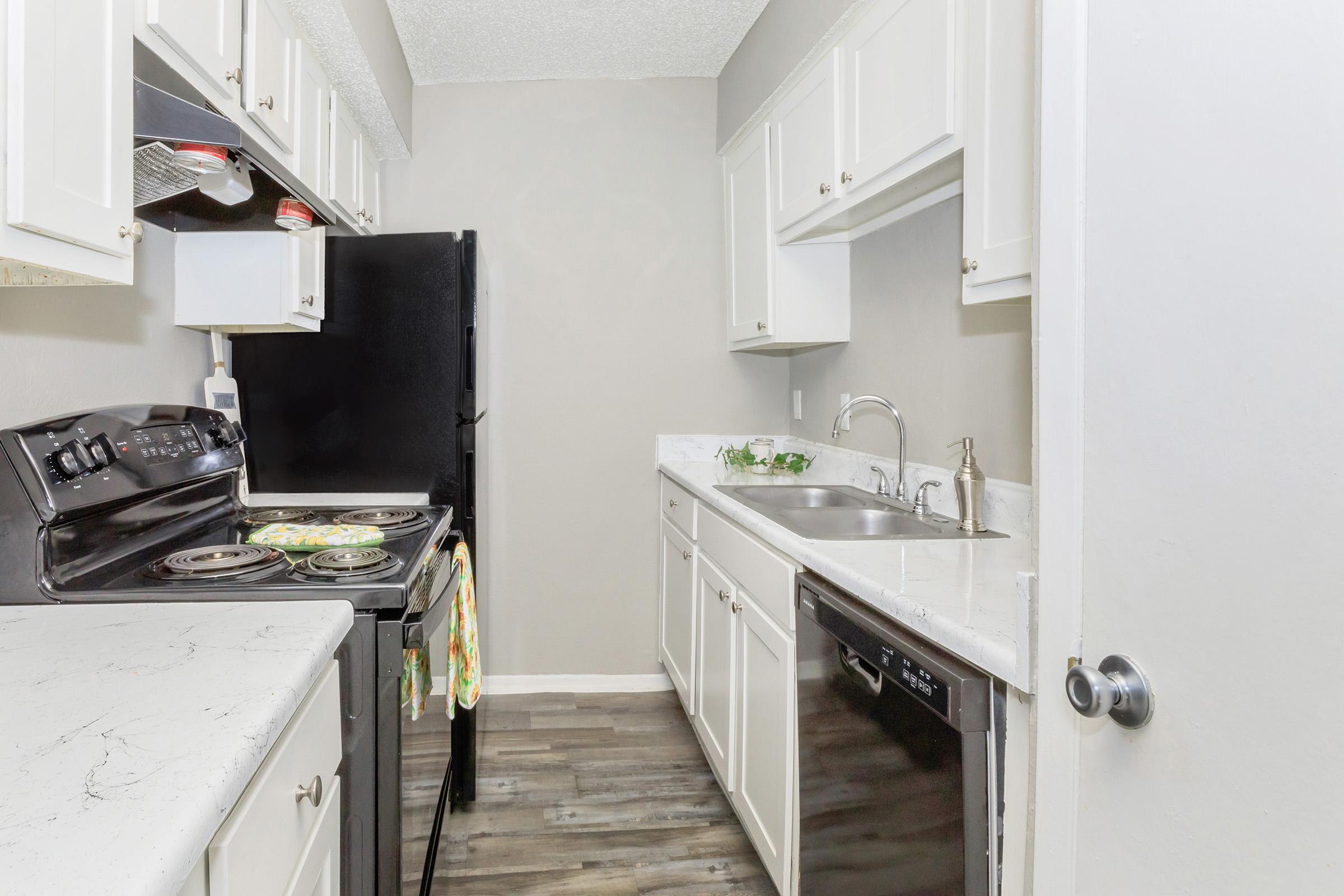 Modern kitchen featuring white cabinetry, a black refrigerator, a stove, and a dishwasher. The countertops are light-colored with a marbled effect, and the floor is a dark wood laminate. Natural light comes in, enhancing the clean and functional space.