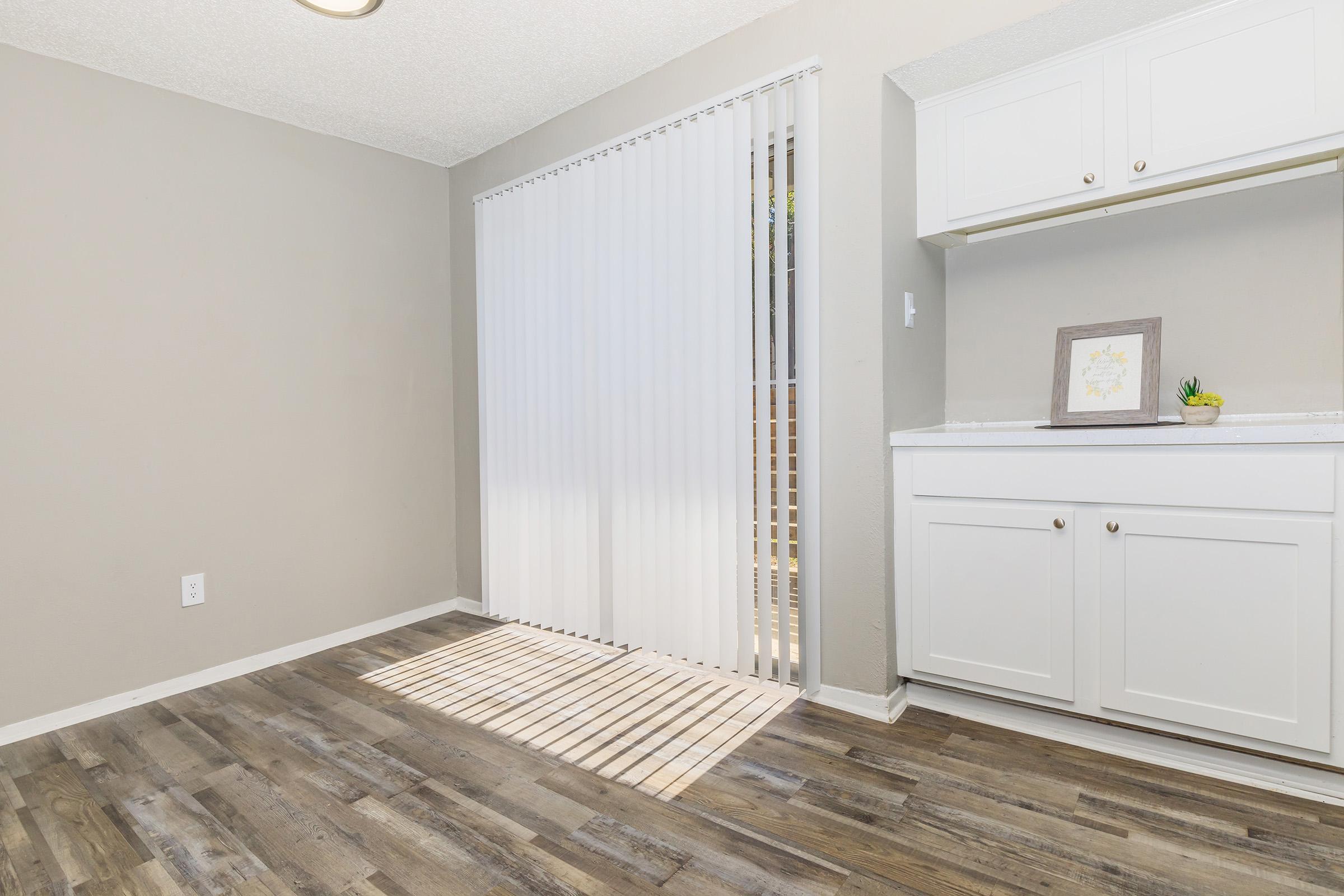 A bright, minimalistic room featuring a light gray wall and wooden flooring. On the left, there are vertical blinds covering a window that lets in natural light, creating a striped pattern on the floor. To the right, there is a white cabinet with a framed picture and a small plant.