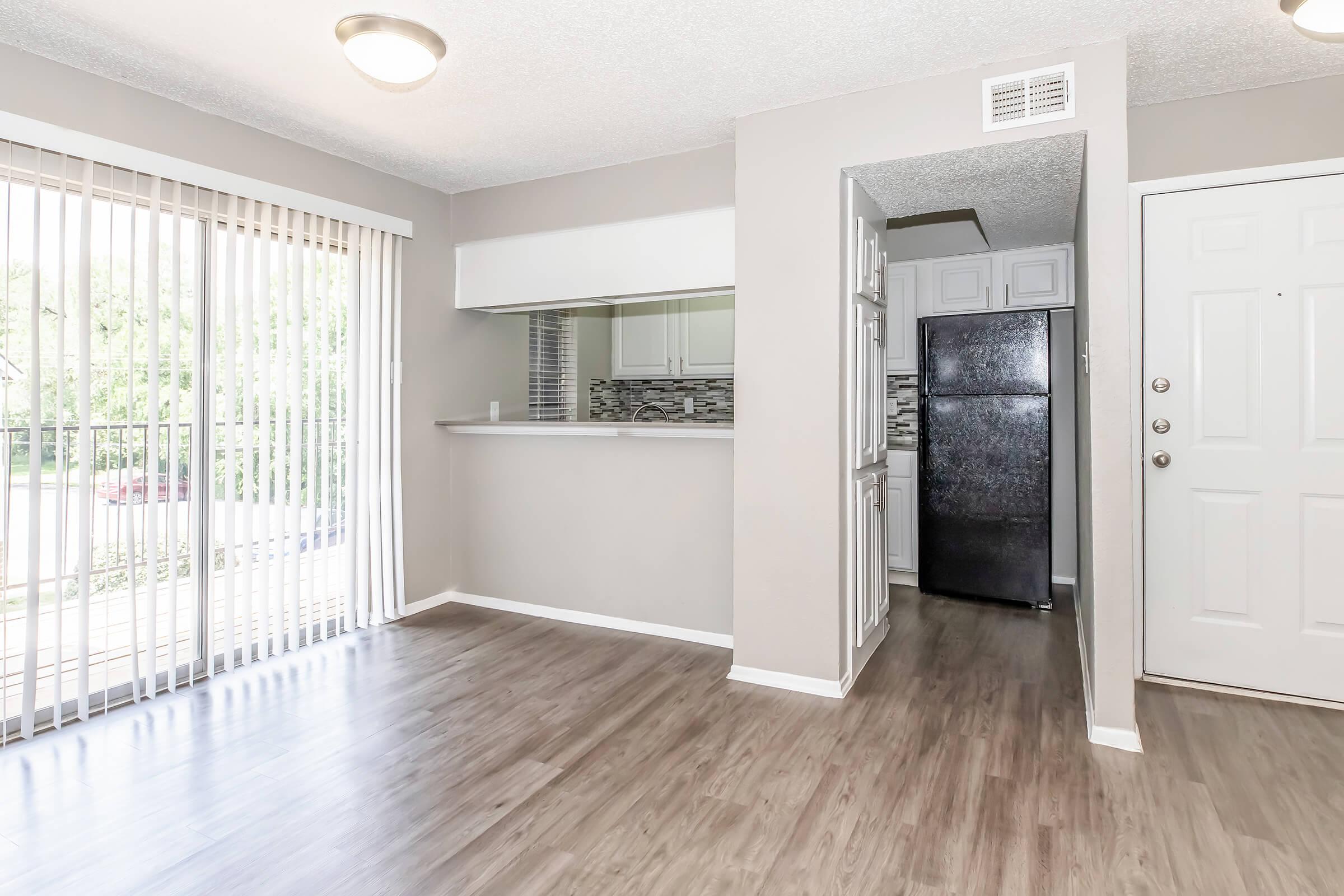 Bright, modern living room featuring light gray walls, wooden flooring, and a sliding glass door that leads outside. A small breakfast bar opens into a kitchen area with black appliances, including a refrigerator. The design is airy and welcoming, perfect for casual gatherings.