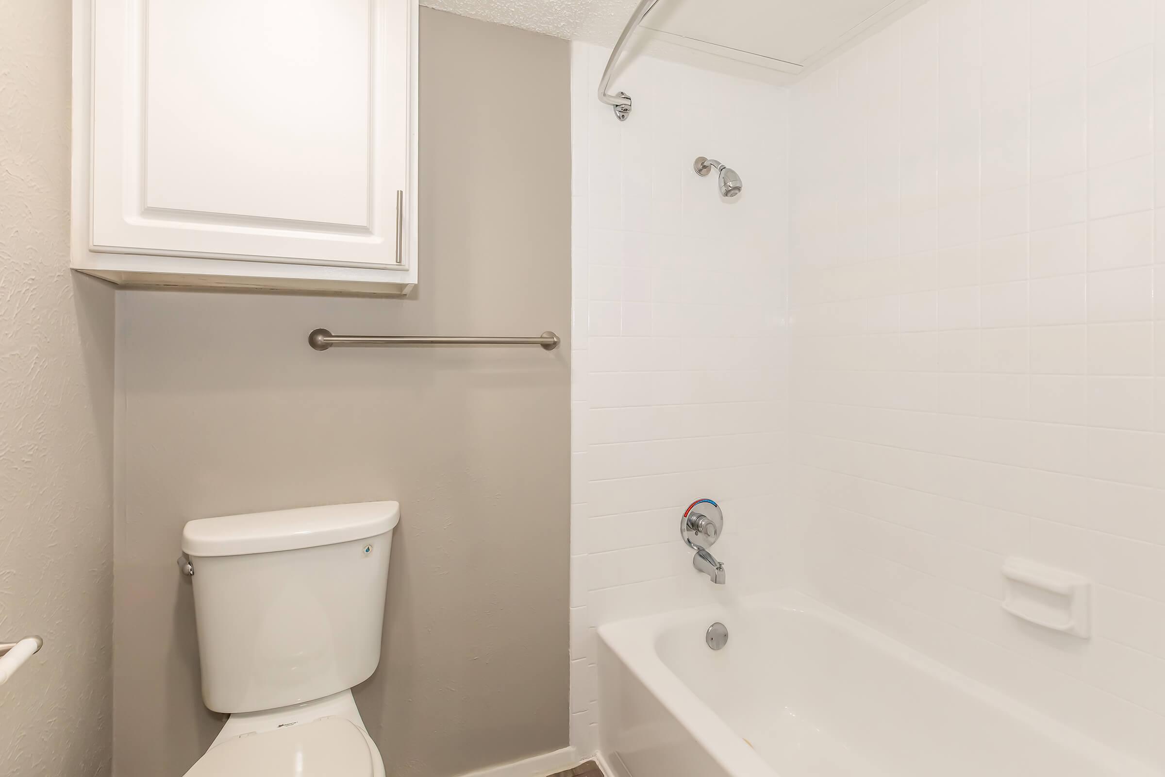 A clean and modern bathroom featuring a white bathtub, a toilet, and a showerhead. The walls are a light gray color, with white tiles surrounding the bathtub. A towel bar is installed on the wall, and there is a white cabinet above the toilet for storage.
