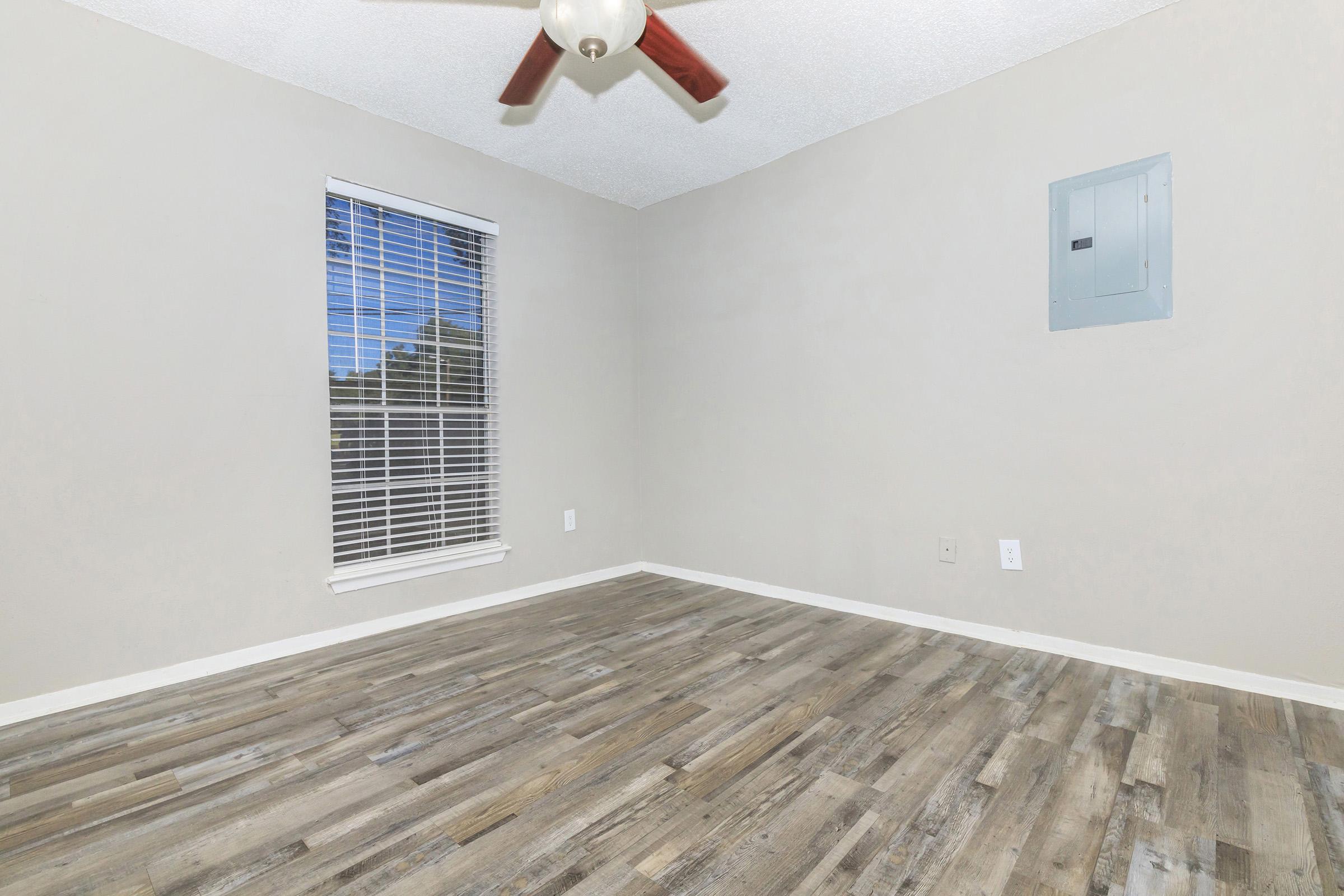 A well-lit, empty room with a light-colored wall, a ceiling fan, a window with blinds, and a laminated wooden floor. An electrical panel is mounted on one wall. The room is devoid of furniture, creating a spacious and open feel.