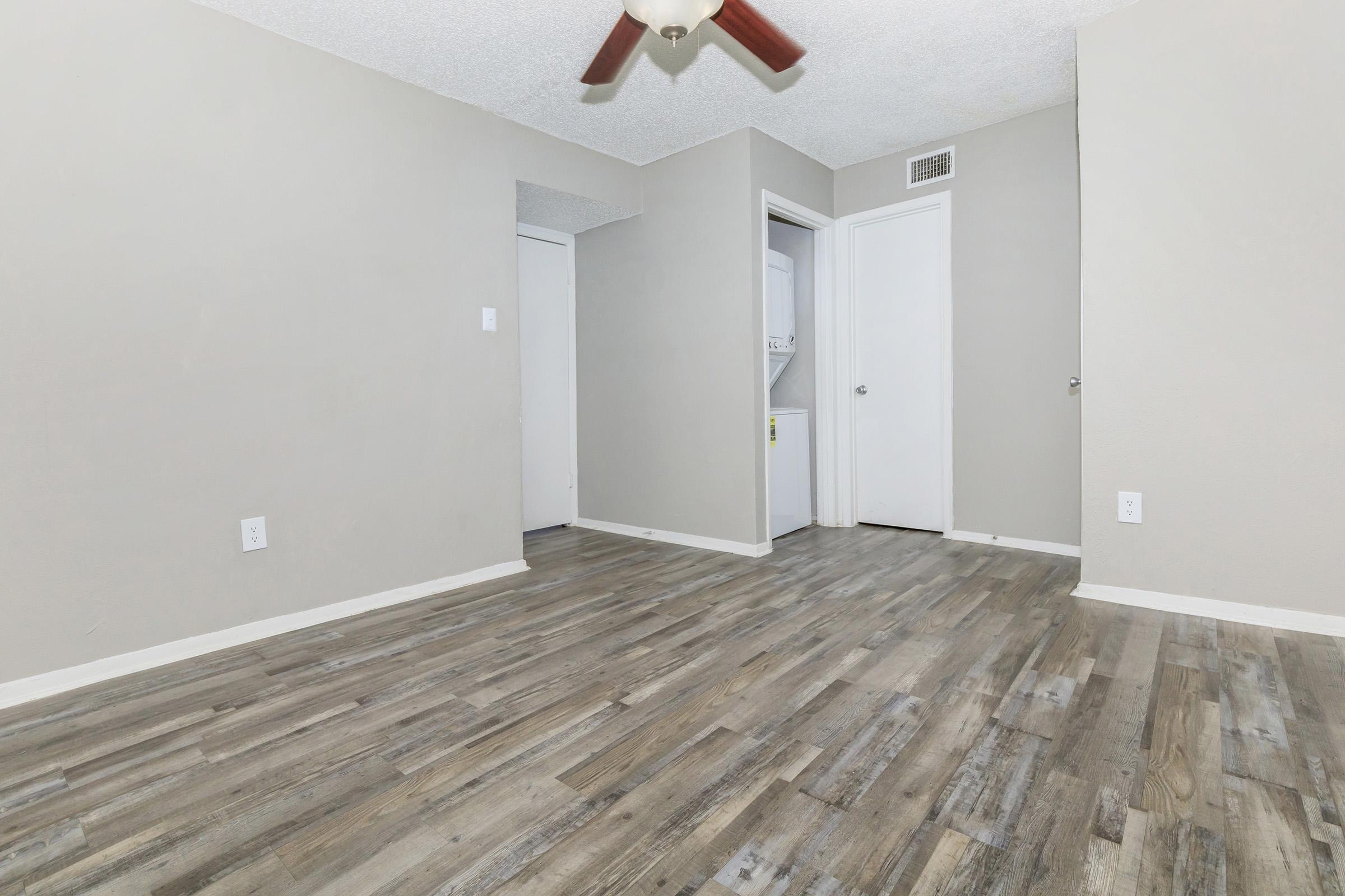 A spacious room featuring light-colored walls and laminate flooring. There's a ceiling fan with wooden blades, and two doorways leading to other areas, one possibly to a laundry room. The overall ambiance is modern and minimalistic, suitable for various uses.