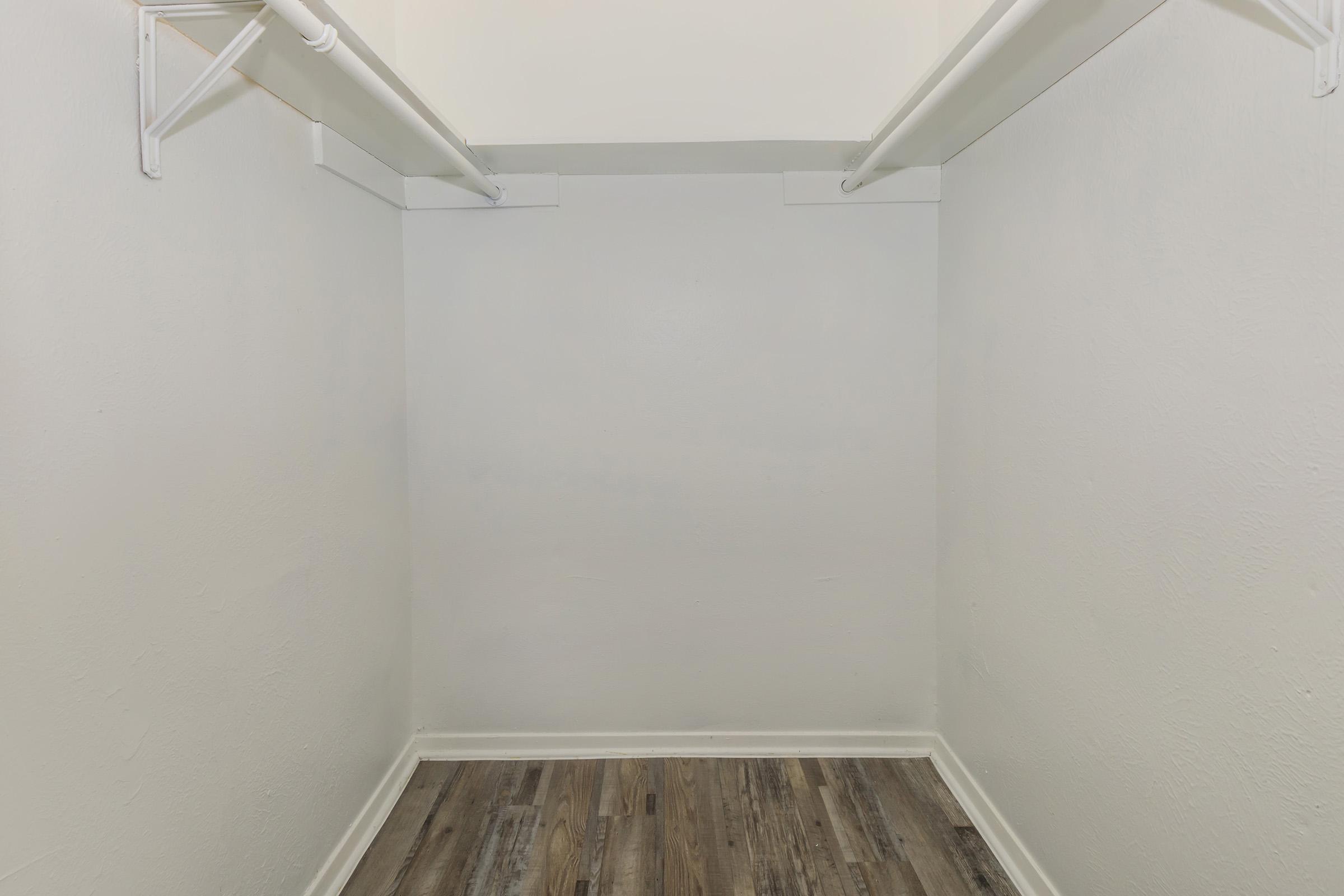 Empty walk-in closet with beige walls and wooden flooring. Two hanging rods on opposite walls with no shelves or storage items visible. The space is well-lit and clean, providing a minimalist appearance.