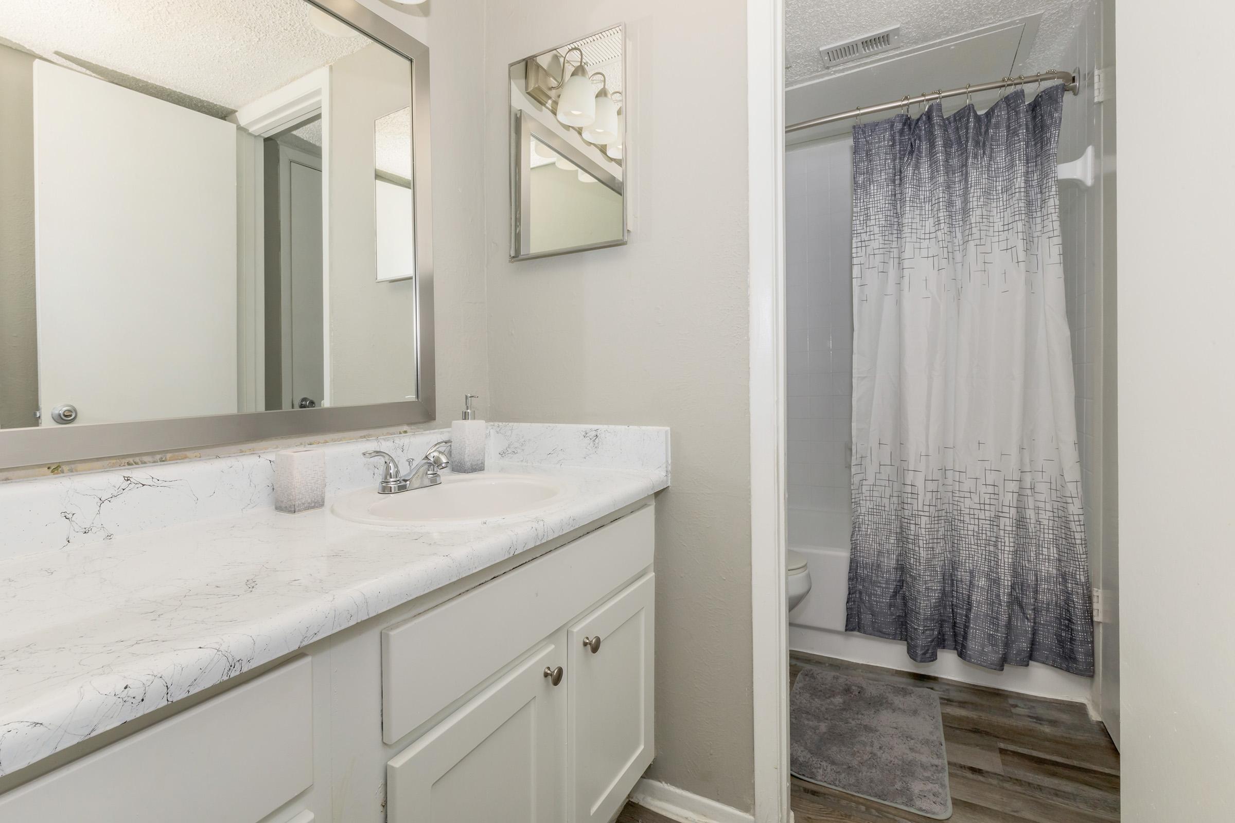 A modern bathroom featuring a white marble countertop with a sink, a decorative mirror, and a shower with a gray and white patterned curtain. The walls are painted light gray, and there is a small bath mat on the floor. The overall design is clean and contemporary.