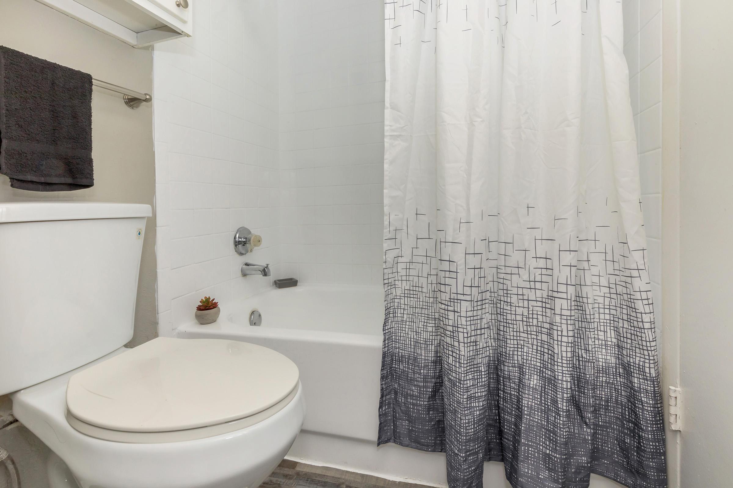 A clean bathroom featuring a white bathtub with a gray patterned shower curtain, a toilet, and a small plant on the tub's edge. The walls are tiled, and there is a towel hanging on a rack. The overall aesthetic is modern and minimalistic.