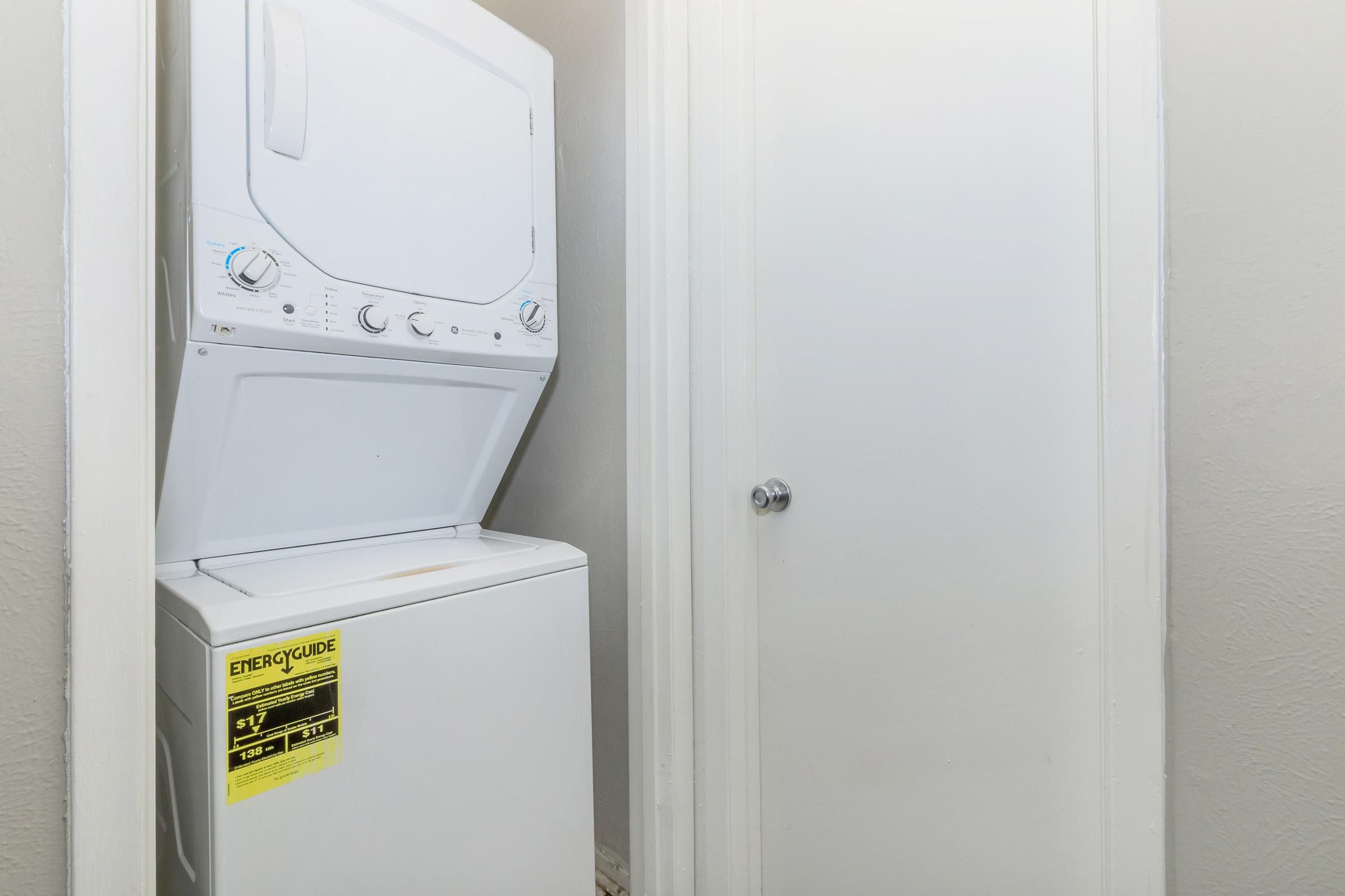 A stacked washer and dryer unit is positioned against a light gray wall. The laundry appliance features control dials on the top section. A closed white door is visible to the right, blending into the wall, suggesting a small laundry area or utility space. A clear energy guide label is attached to the dryer.