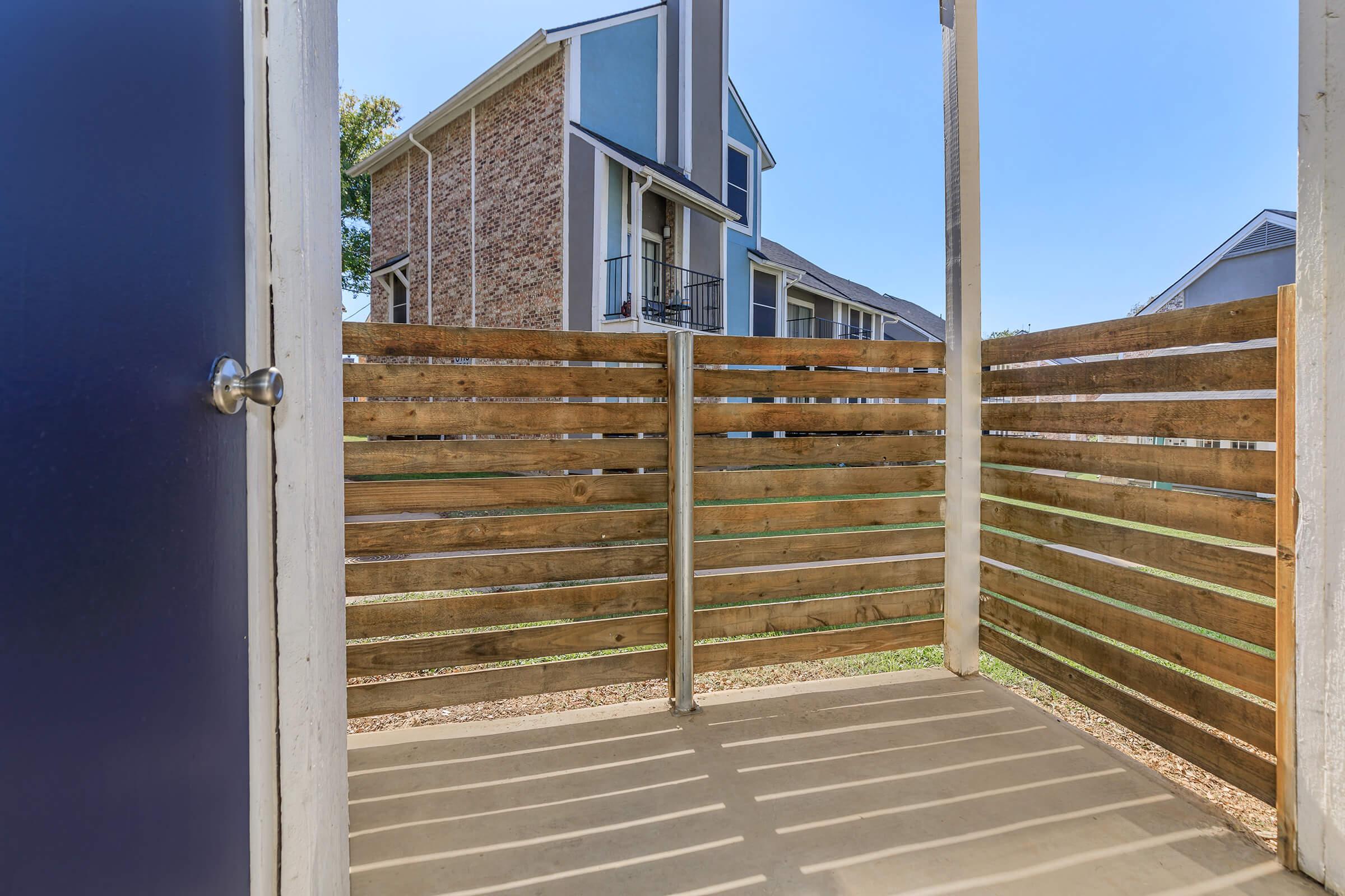 View of a wooden balcony with vertical slats, looking out toward a brick building with a balcony. The sky is clear and bright, casting shadows on the floor of the balcony. The setting appears to be a residential area with a well-maintained outdoor space.