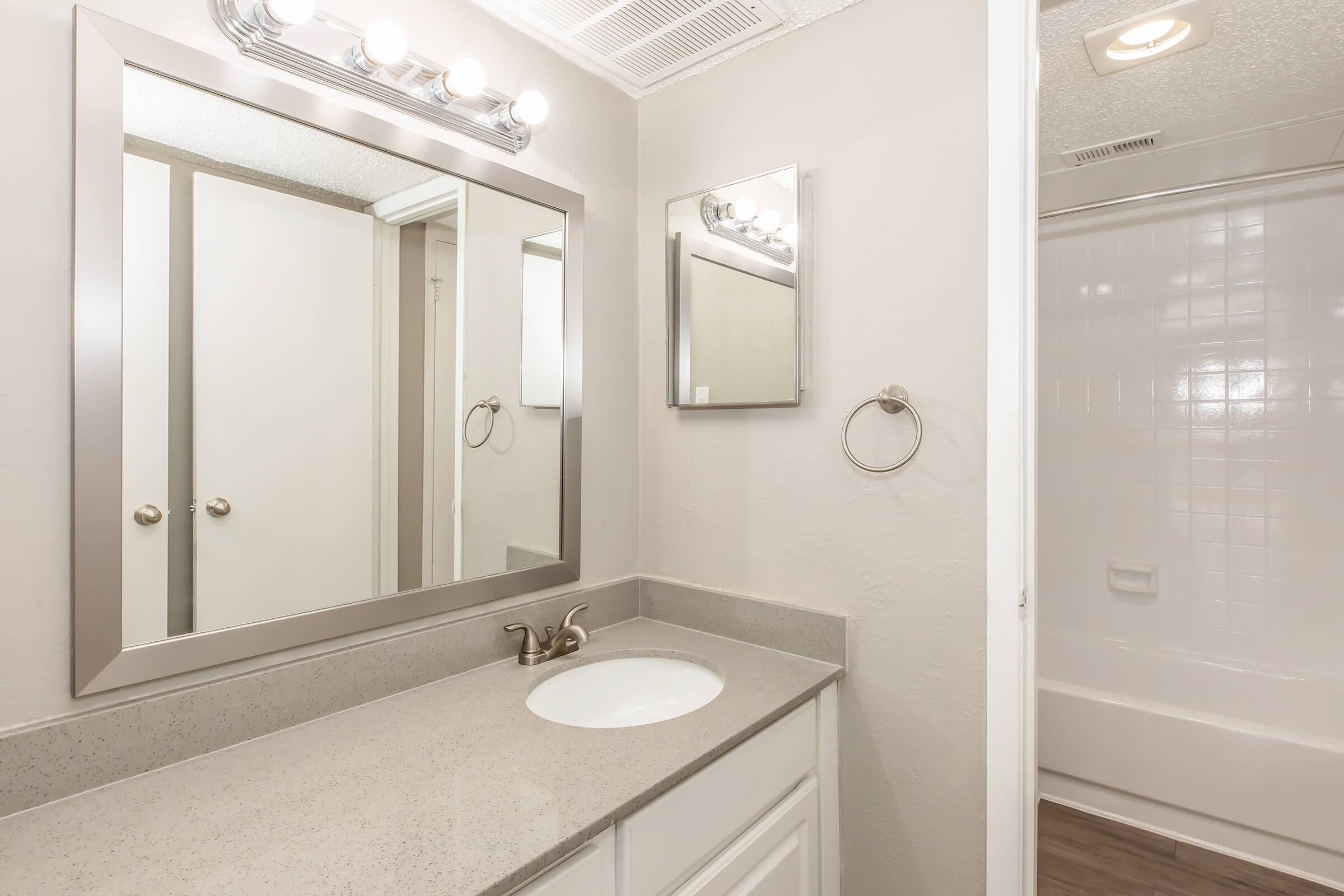 A modern bathroom featuring a double sink vanity with a smooth countertop, two mirrors above the sinks, and a towel ring on the wall. The room is painted in light gray, with a tub and shower area visible through an open door, enhancing the spacious feel of the design.