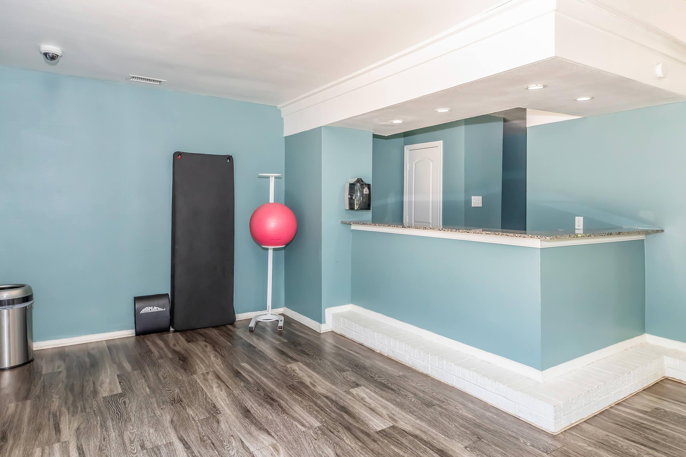 A spacious room with light blue walls, featuring a small reception counter, a large red exercise ball on a stand, a black exercise mat, a trash can, and a white tiled area. The floor is made of dark wood, creating a modern and clean aesthetic.