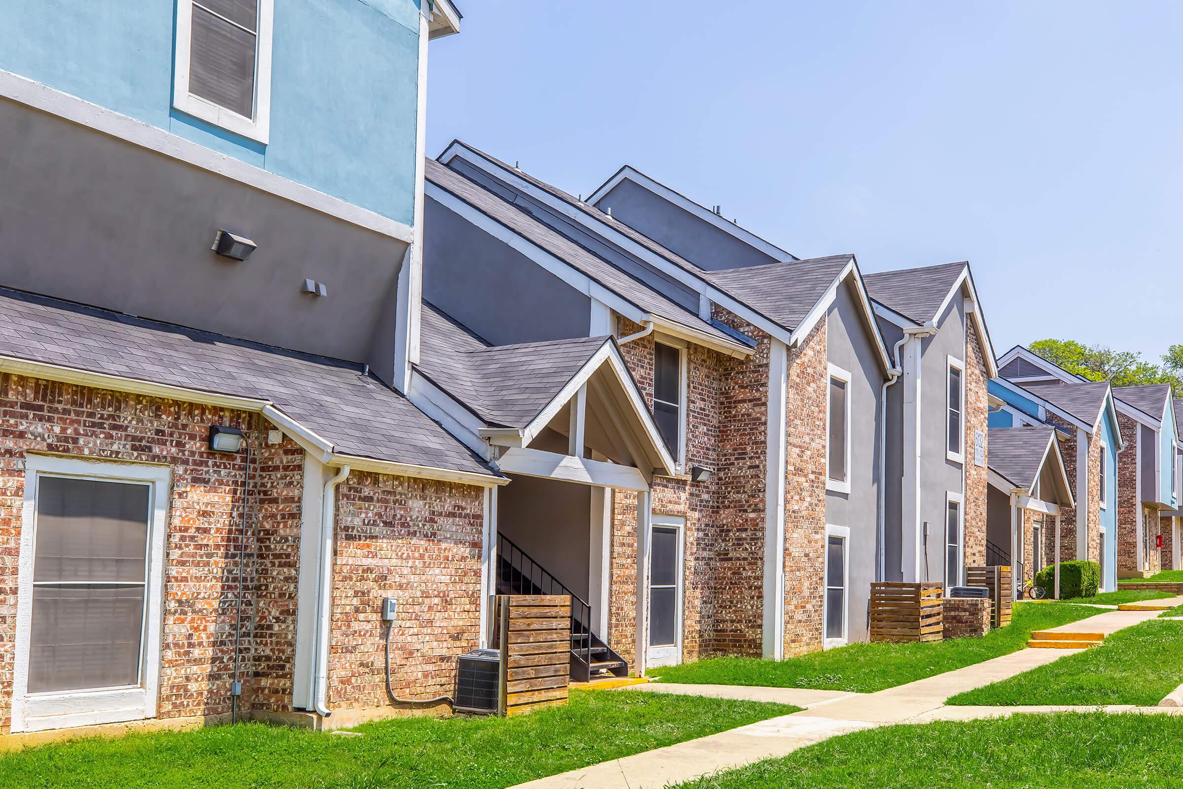 A row of modern townhouses with a mix of brick and painted exteriors. The buildings have multiple levels, with sloped roofs and grassy landscaping in front. Bright blue skies are visible above, creating a sunny atmosphere. Pathways lead to the entrances of the homes.