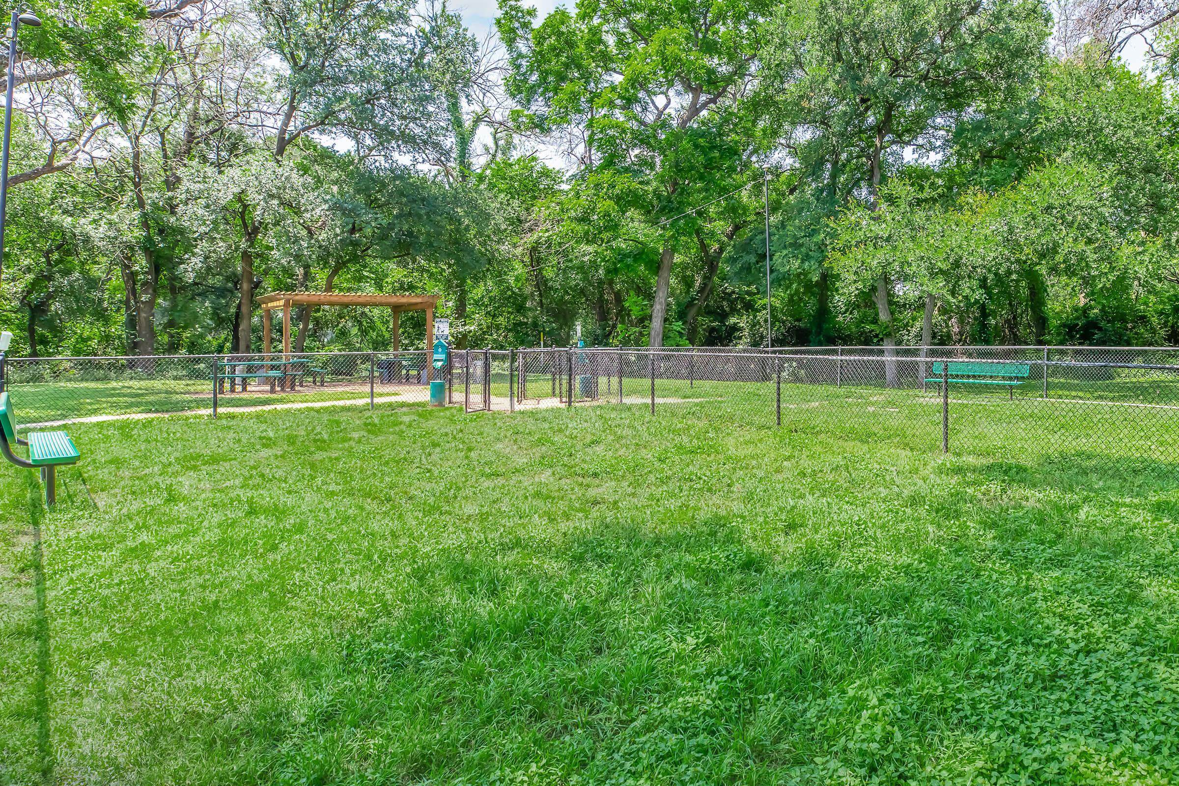 A spacious grassy area enclosed by a fence, featuring a dog park with a gazebo, picnic tables, and benches under the shade of trees. The scene is bright and sunny, showcasing the greenery and outdoor space designed for dogs and their owners.