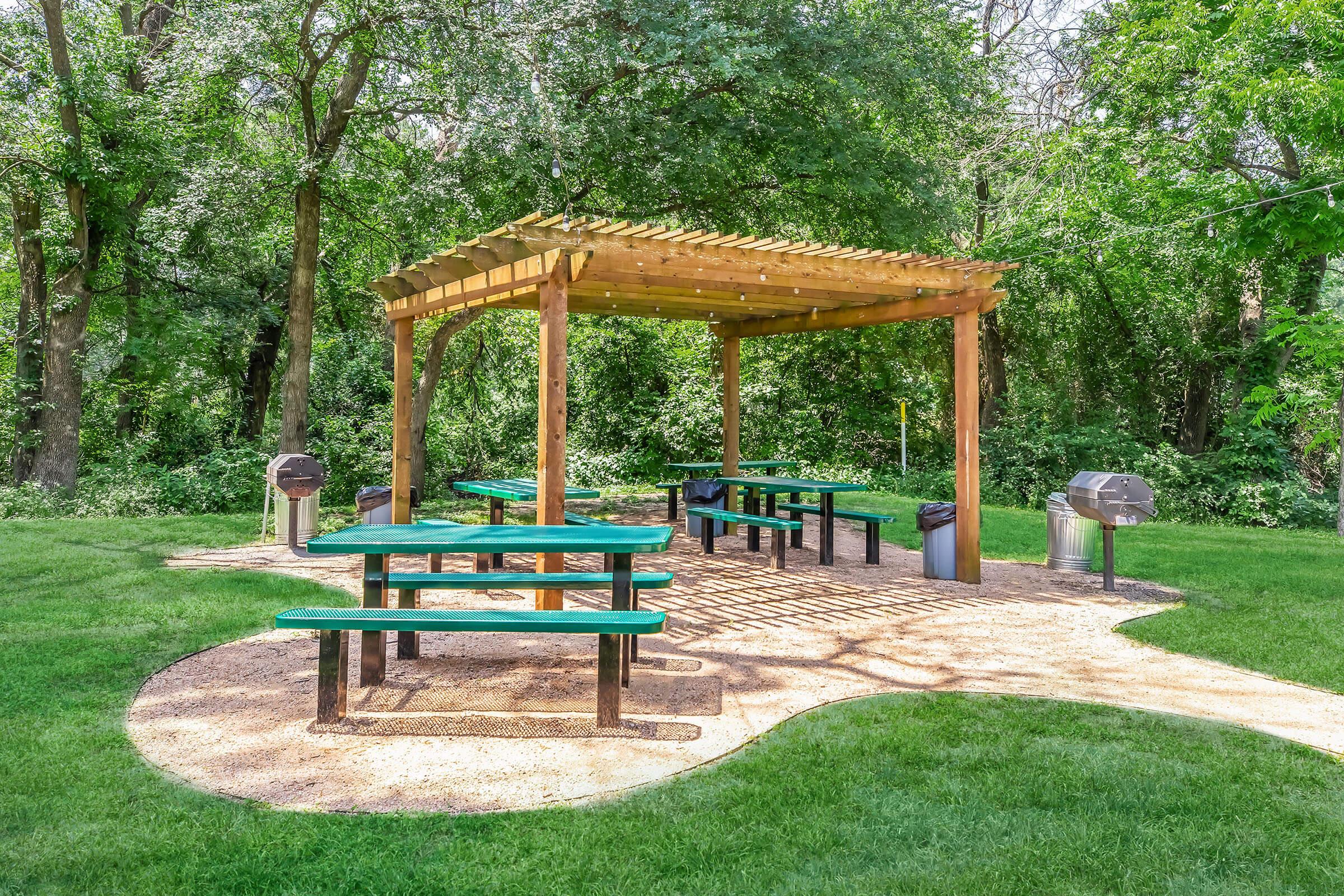 A shaded picnic area featuring green tables and benches under a wooden pergola, surrounded by grassy space and trees. There are two grills positioned nearby and a path leading to the seating area, creating an inviting spot for outdoor gatherings.