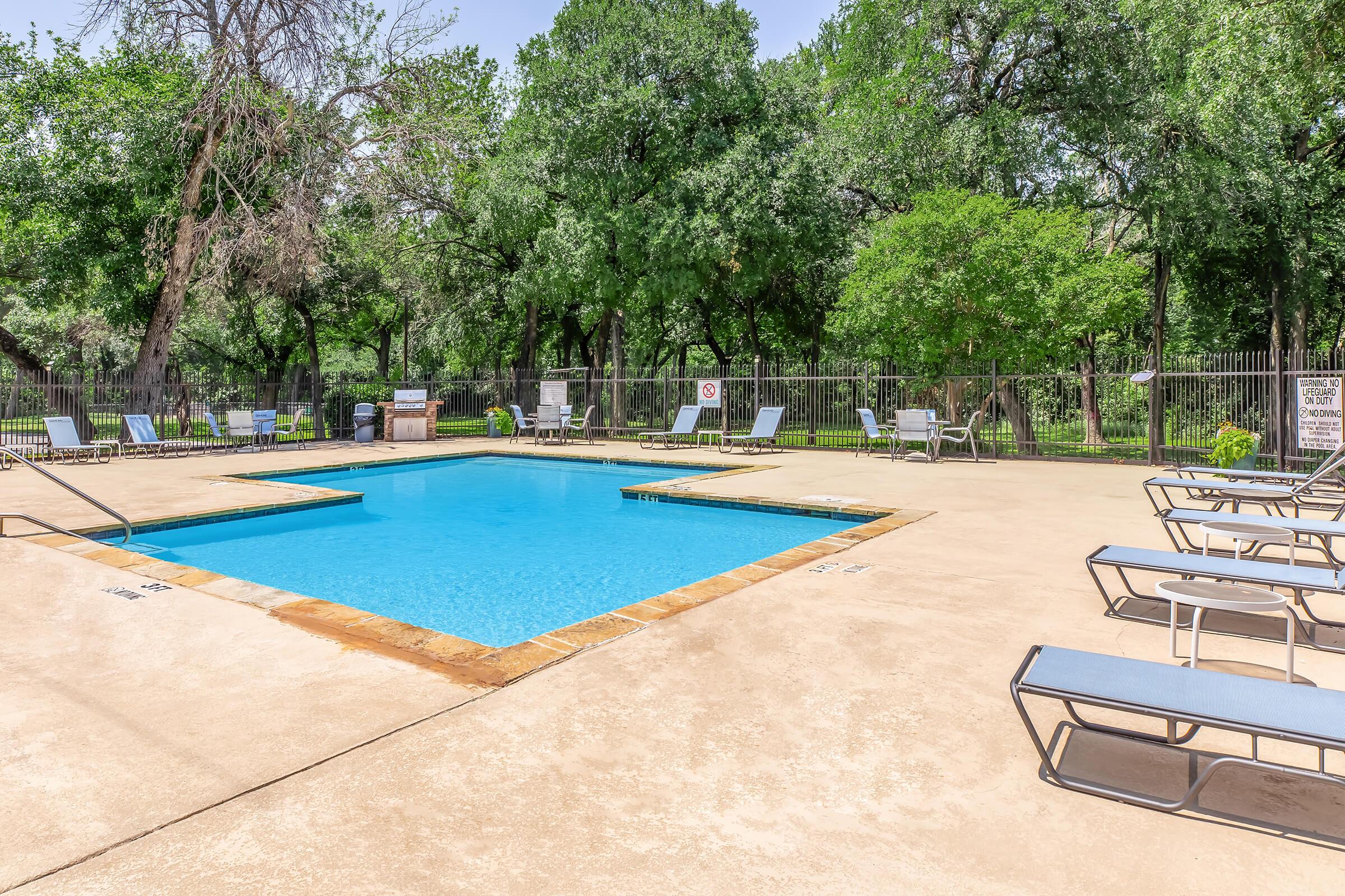 A clear swimming pool surrounded by a concrete deck with lounge chairs. Lush green trees and foliage line the background, providing a serene atmosphere. A barbeque area is visible nearby, along with safety signs and a gated fence. The scene is bright and inviting, perfect for relaxation and leisure.