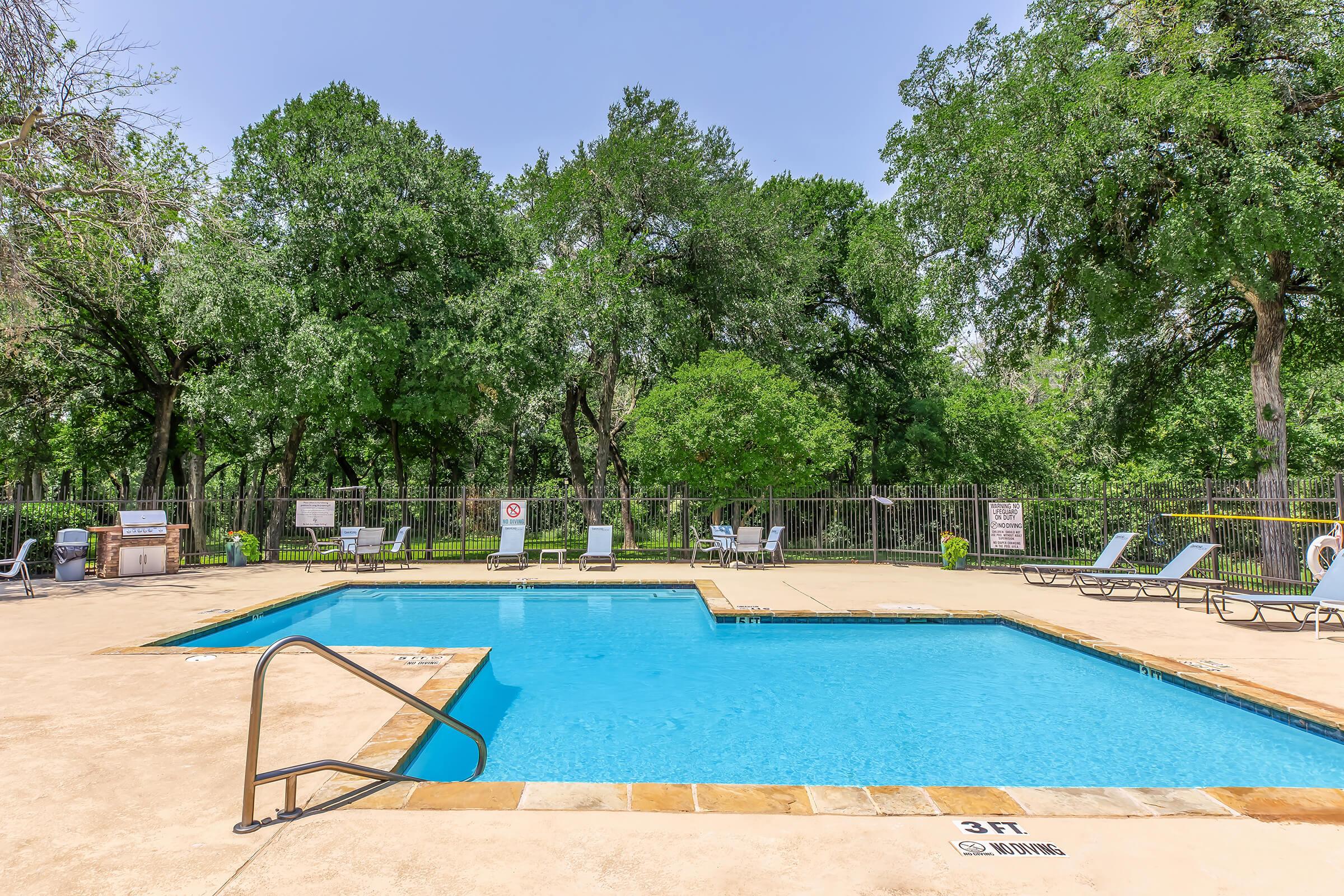 A clear blue swimming pool surrounded by lounge chairs and greenery. The pool area features a shallow end marked with a sign indicating "3 ft." and a fenced perimeter with trees in the background, creating a tranquil outdoor setting.