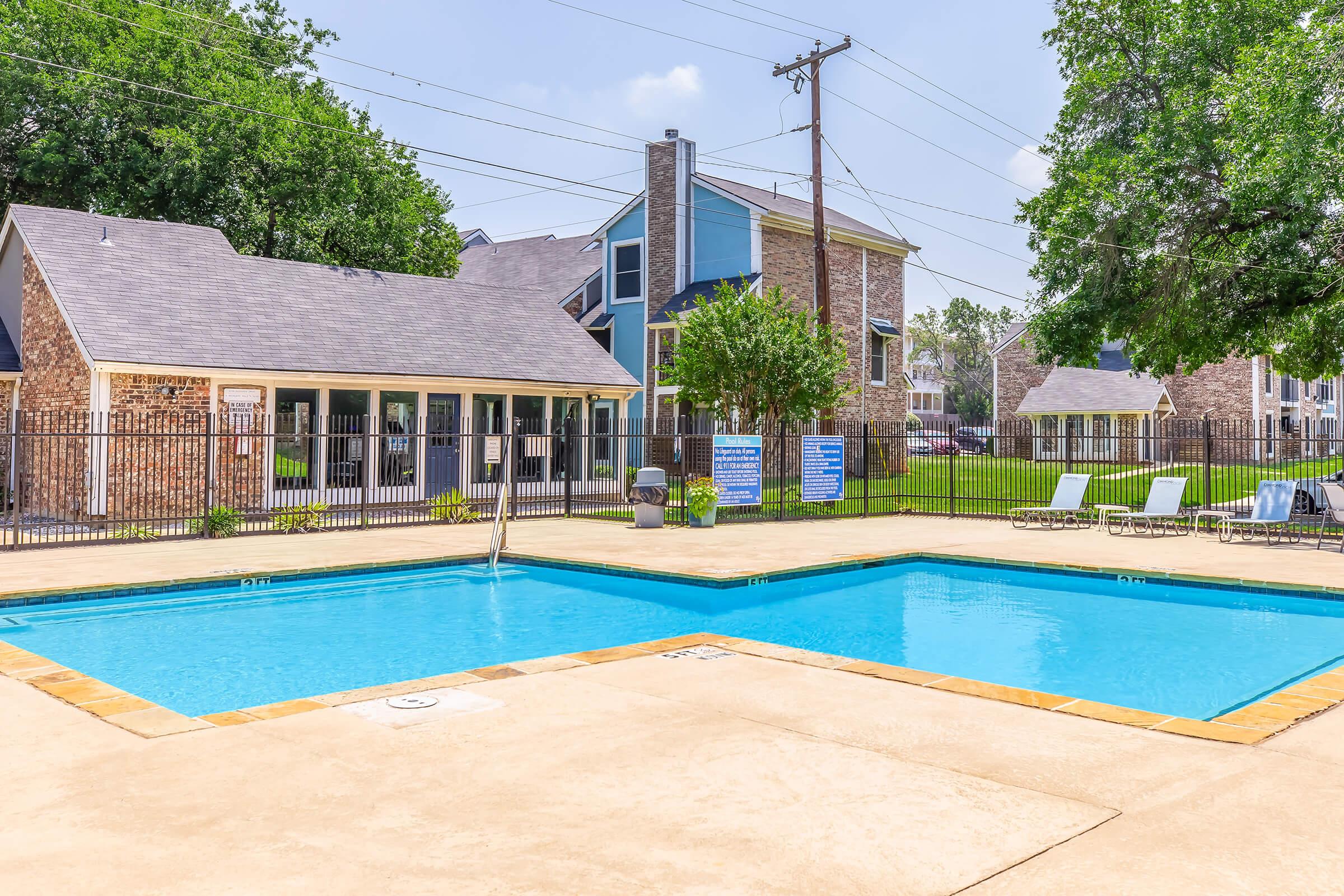 A bright and inviting outdoor swimming pool surrounded by a tan concrete deck, lounge chairs, and a fenced area. In the background, there are several buildings with a mix of brick and siding, green trees, and a clear blue sky overhead. The atmosphere is relaxed and perfect for leisure activities.