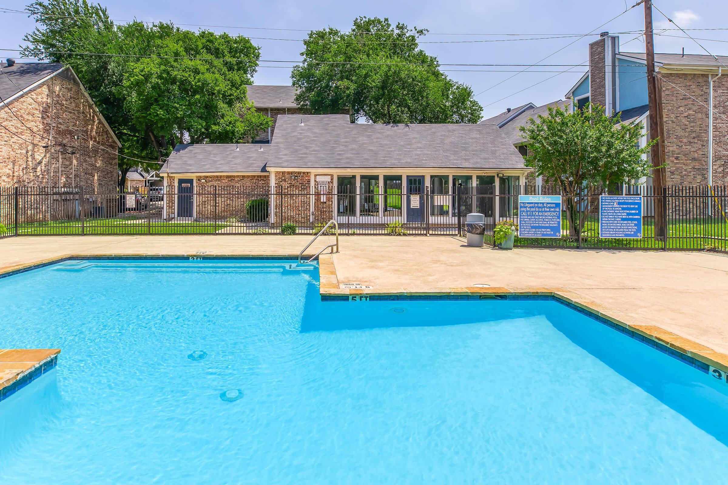 A clear blue swimming pool with a shallow area, surrounded by concrete decking. In the background, there is a brick building with large glass doors and several trees. The area is enclosed by a black metal fence, and additional signage is visible on the fence near the pool.