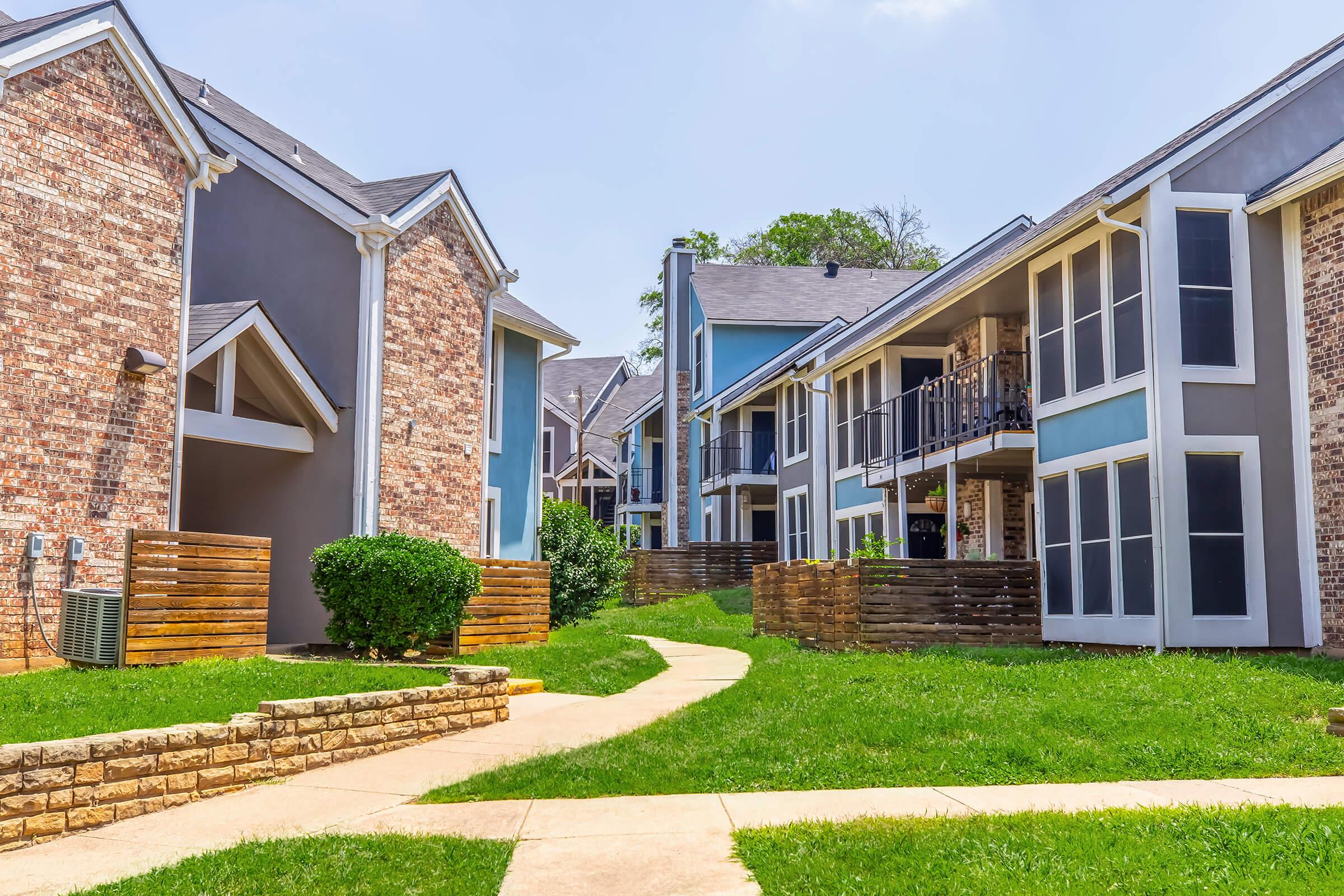 A view of a residential complex featuring modern buildings with brick and blue exteriors. A paved walkway winds through well-maintained green lawns, bordered by shrubs and wooden fences. The scene is under a clear blue sky, showcasing a pleasant and inviting living environment.