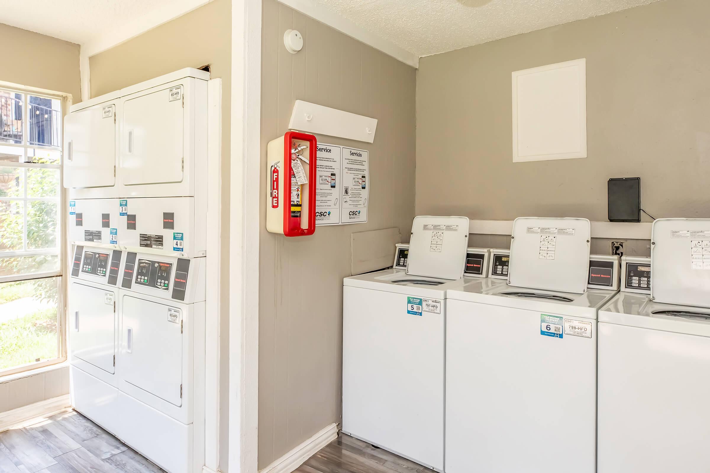 A clean and organized laundry room featuring stacked washer and dryer units on the left and multiple traditional washing machines and dryers on the right. A fire extinguisher is mounted on the wall, and large windows allow natural light to brighten the space.