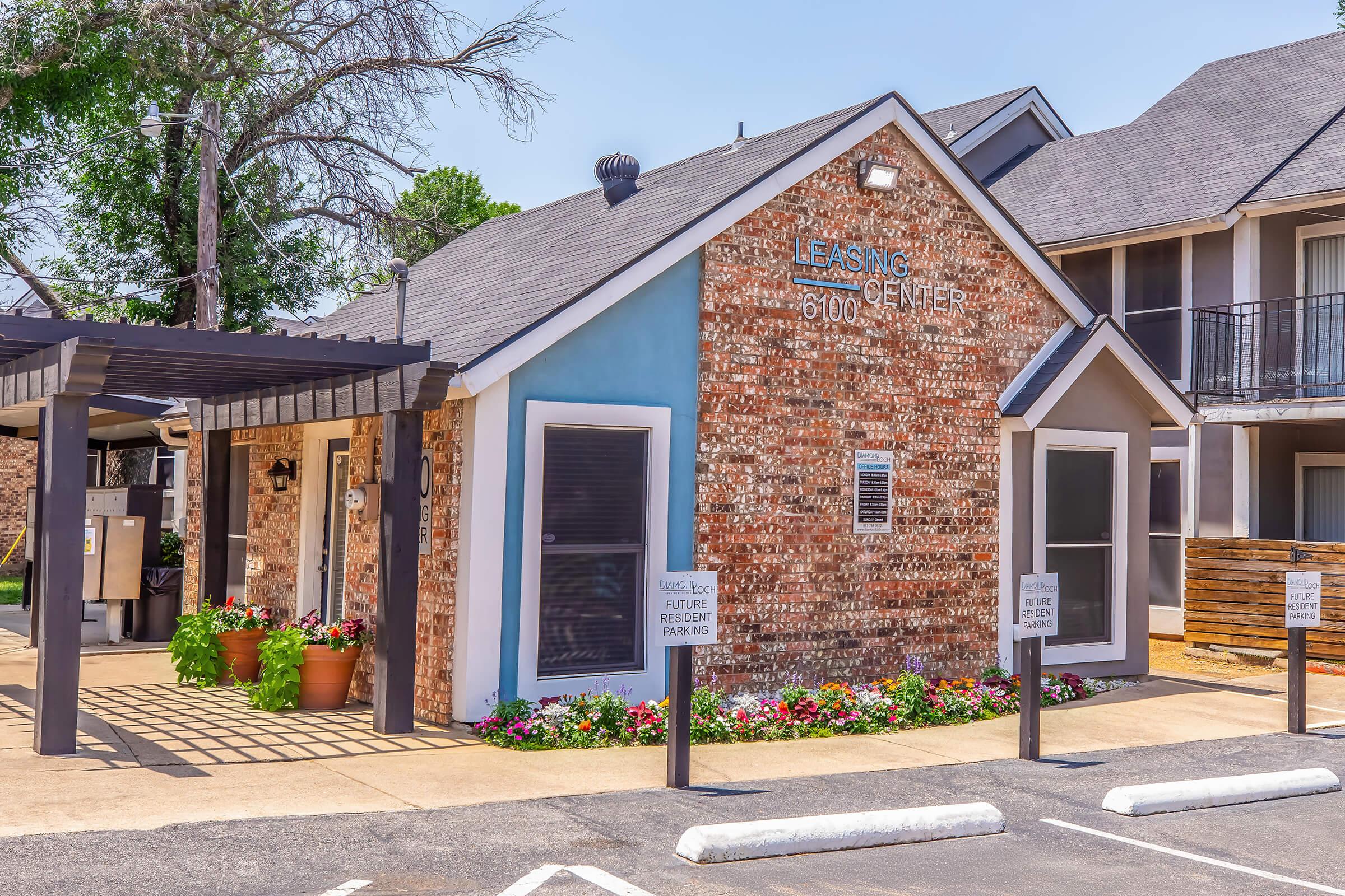 Leasing center of an apartment complex featuring a brick facade, blue accent wall, and flower beds in front. Signs indicate leasing information, and adjacent buildings can be seen in the background under a clear blue sky.