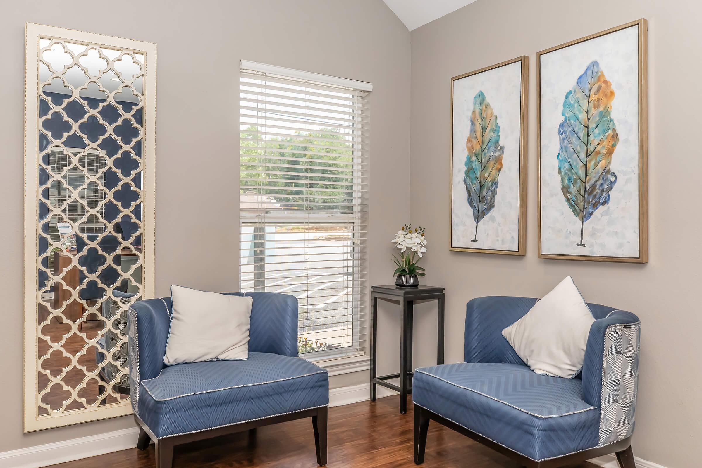 A cozy sitting area featuring two blue armchairs with white pillows, a small black side table, and a floral arrangement. The walls are adorned with two framed artworks of leaves in shades of blue and orange. A decorative mirror with an intricate design hangs beside a window with blinds, allowing natural light to enter.
