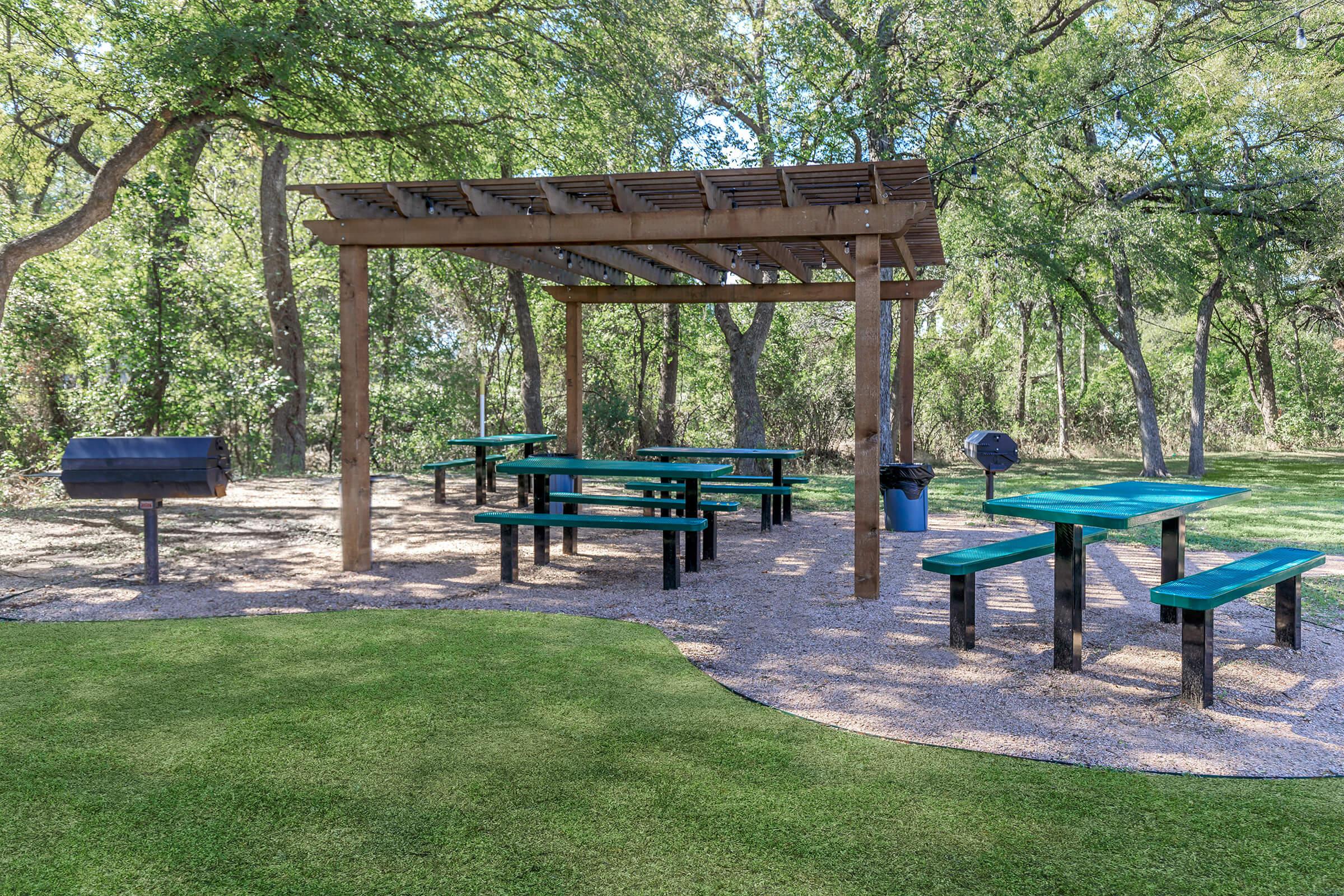 A covered picnic area featuring multiple green tables and benches, set on a grassy surface among trees. A charcoal grill is visible nearby, creating a suitable space for outdoor gatherings and barbecues.