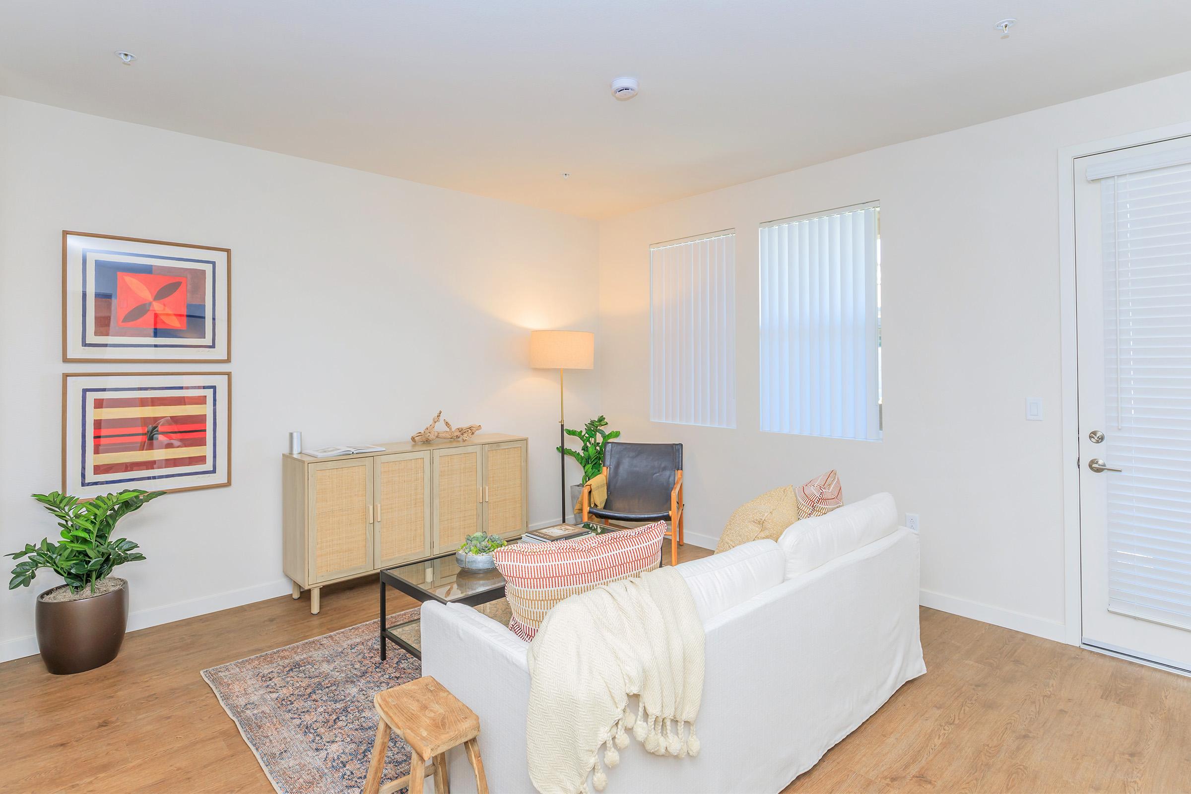 A cozy living room featuring a light-colored sofa adorned with a throw blanket, a modern armchair, and a wooden sideboard. Two framed artworks hang on the wall, and there's a potted plant in the corner. Natural light streams in through partially open blinds, creating a welcoming atmosphere.