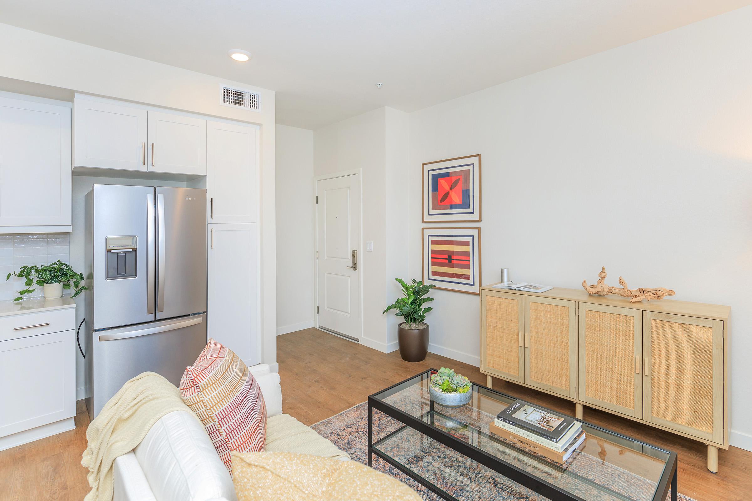 A cozy living room area featuring a white sofa with colorful pillows, a glass coffee table with books, a beige cabinet, a green plant in a pot, and framed artwork on the walls. The kitchen area is visible with modern appliances, including a stainless steel refrigerator. Warm, inviting decor enhances the space.