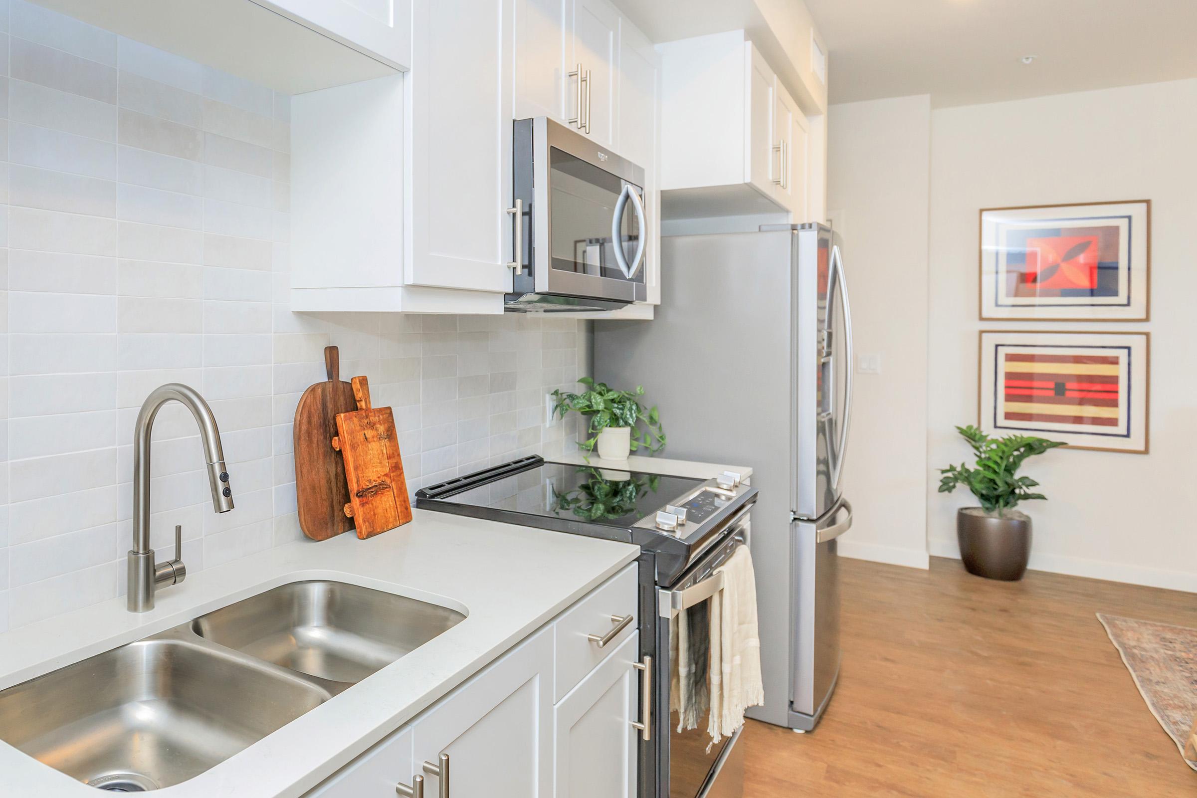 Modern kitchen featuring white cabinetry, stainless steel appliances, and a double sink. A wooden cutting board rests on the countertop, alongside a decorative plant. The floor is wooden, and in the background, there are framed artworks hanging on the wall.