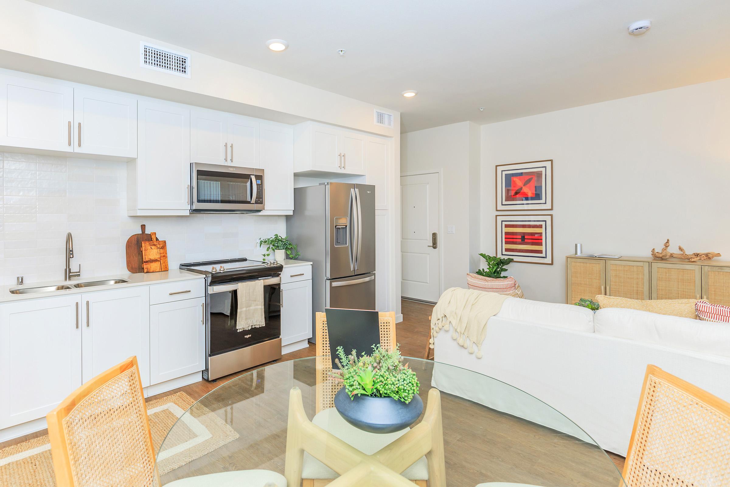 Modern kitchen and living area in a bright, open-concept apartment. The kitchen features white cabinets, stainless steel appliances, and a small wooden accent piece. The living area has a light-colored sofa, a decorative rug, and a plant on a dining table surrounded by wooden chairs. Wall art adds color and personality.