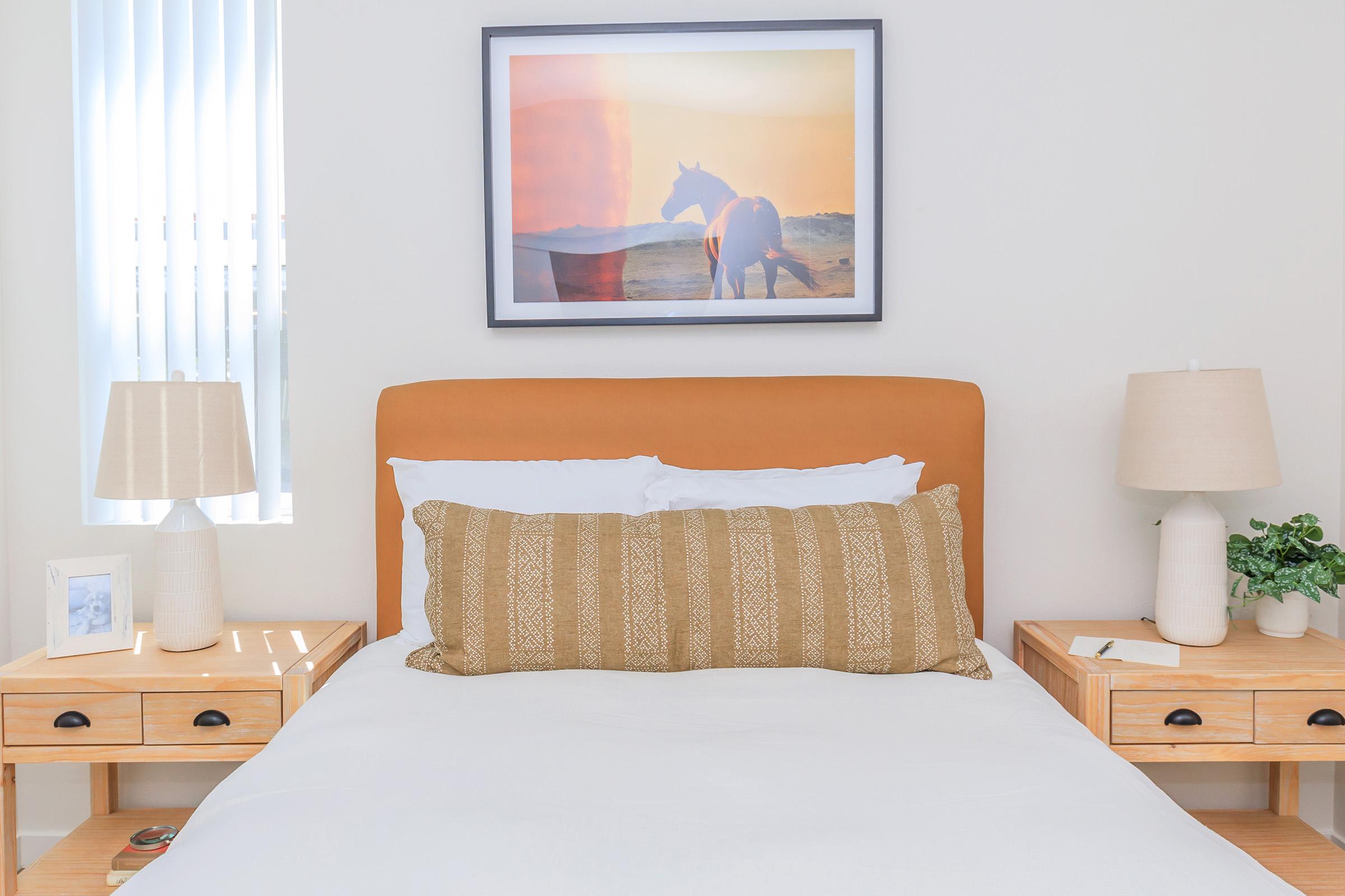 A serene bedroom scene featuring a bed with white bedding and a large decorative pillow in shades of brown. The headboard is orange. On either side of the bed are wooden nightstands with beige lamps and a small potted plant. A framed picture of a horse against a sunset hangs above the bed. Natural light filters through the window.