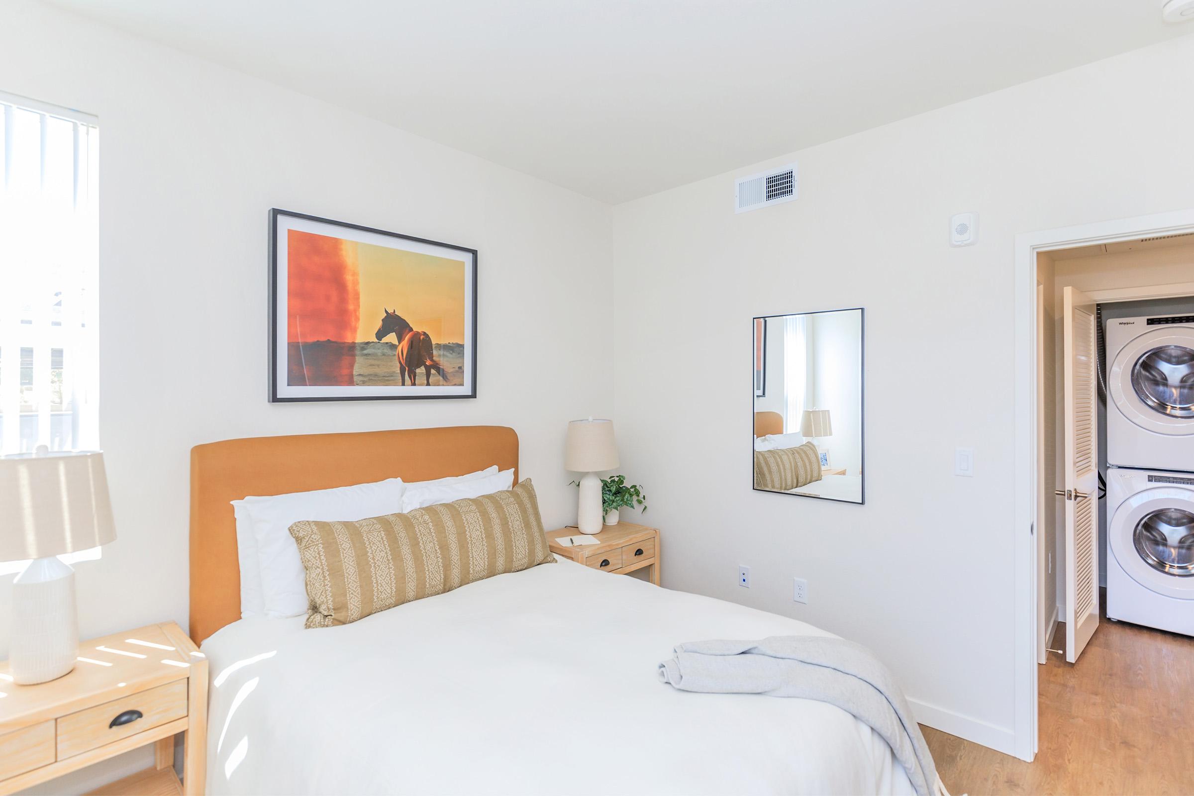 A bright and cozy bedroom featuring a queen-sized bed with white linens and a decorative beige pillow. There's a framed horse painting on the wall above the bed, a bedside table with a lamp, and a full-length mirror. An open door reveals a laundry area with stacked washer and dryer in the background.