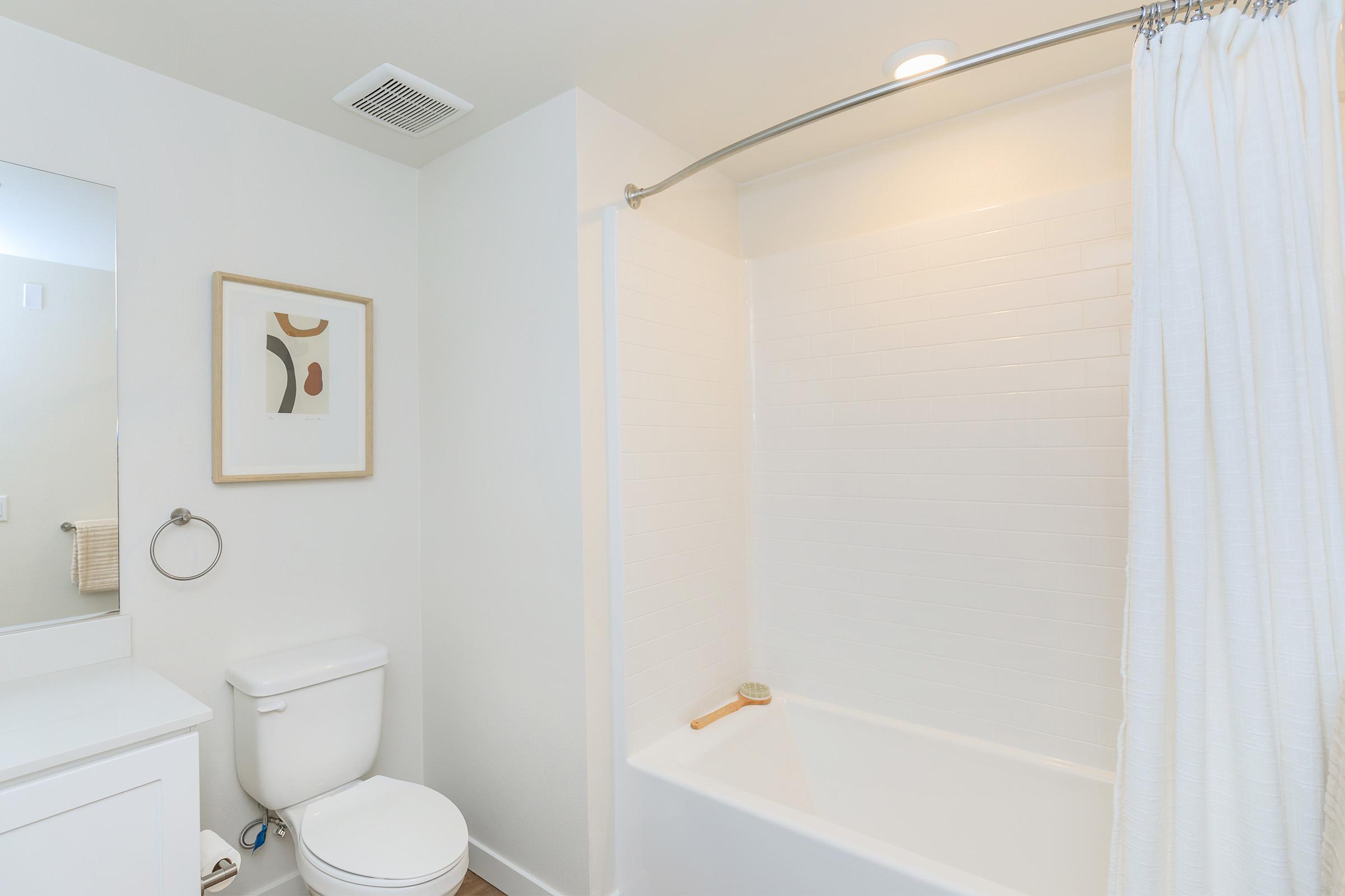 A clean and modern bathroom featuring a white tiled bathtub with a shower curtain, a toilet, a minimalistic sink with a mirror above, and a framed abstract art piece on the wall, accentuated by soft lighting. The overall aesthetic is bright and contemporary.