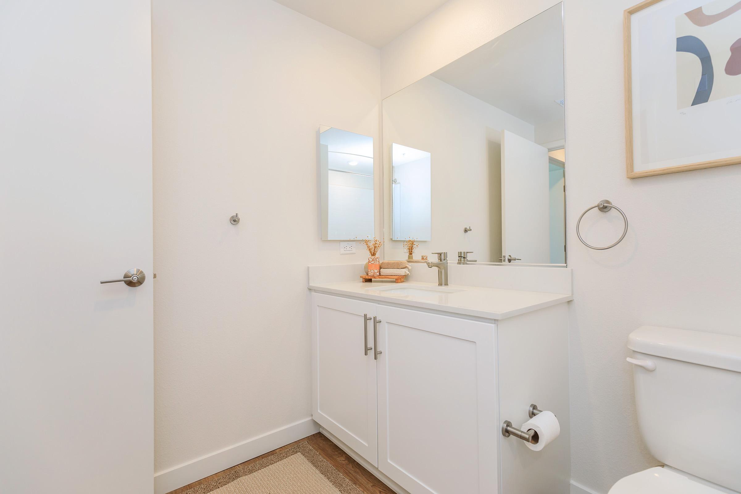 A modern bathroom featuring a white vanity with two mirrors, a toilet, and a towel holder. The space is bright and minimalistic, with light-colored walls and a small decorative item on the countertop. A beige rug is laid on the floor, adding warmth to the clean design.