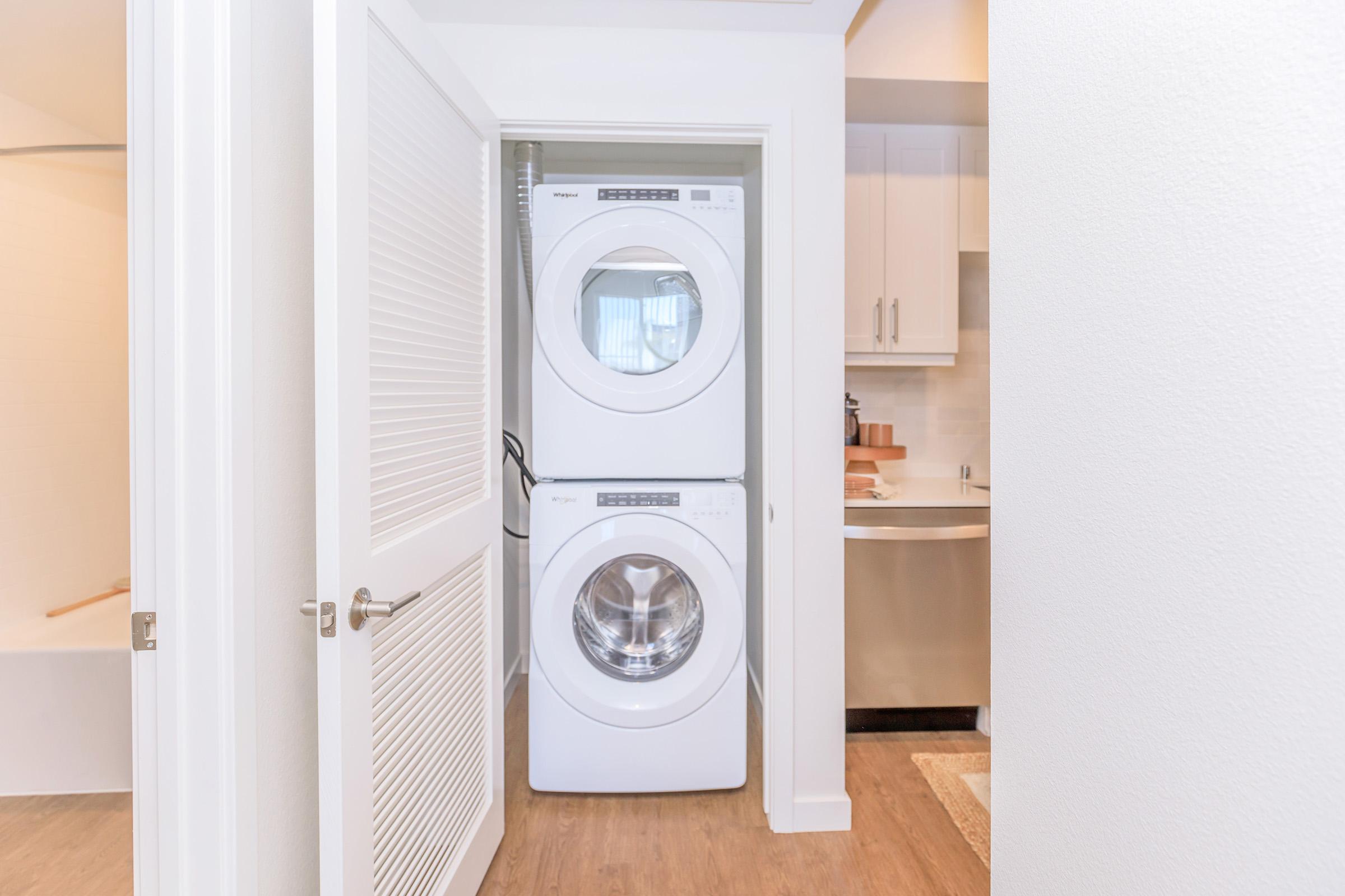 A stacked washer and dryer unit inside a small laundry closet, with light-colored walls and wooden flooring. Visible is part of a kitchen area in the background, featuring white cabinetry and a countertop. The door to the closet is partially open.