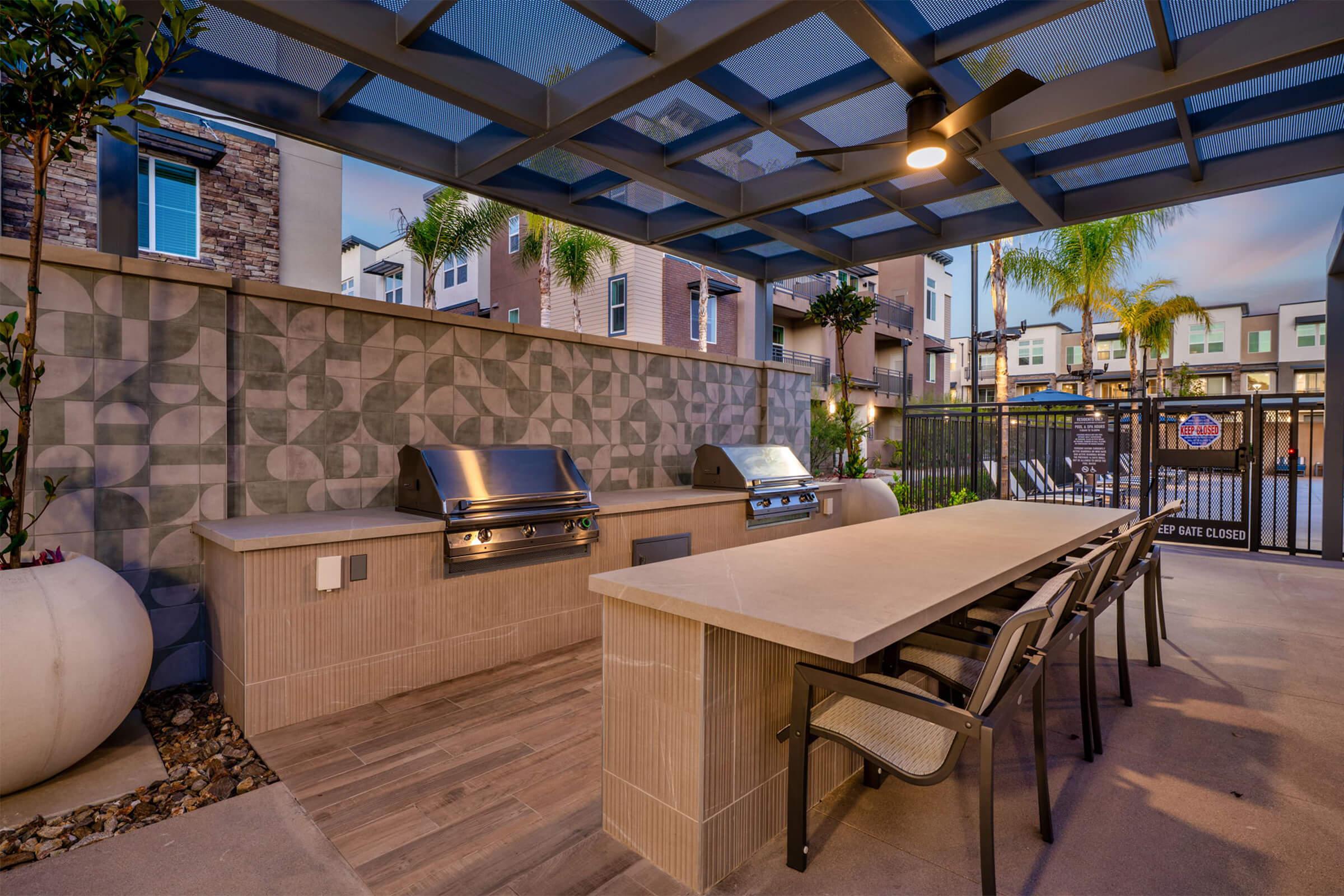 Outdoor BBQ area featuring a modern design with two stainless steel grills, a long stone table, and several chairs. The space is covered with a pergola, surrounded by decorative plants, and offers a stylish atmosphere for outdoor cooking and dining.
