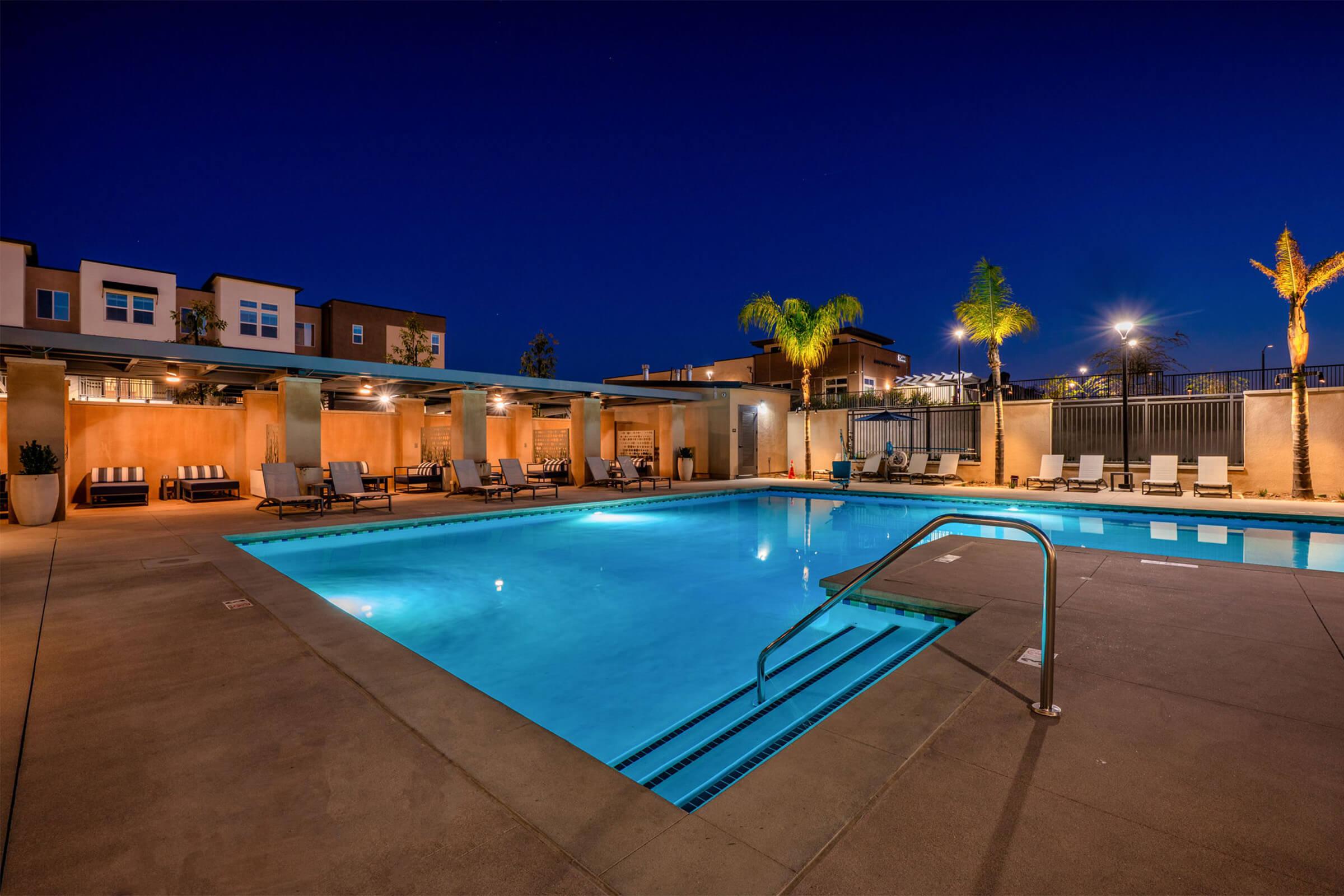 A serene nighttime view of a swimming pool surrounded by lounge chairs and palm trees. The pool’s water is illuminated, reflecting the surrounding lights, with a modern building visible in the background. The atmosphere is calm and inviting, perfect for relaxation.