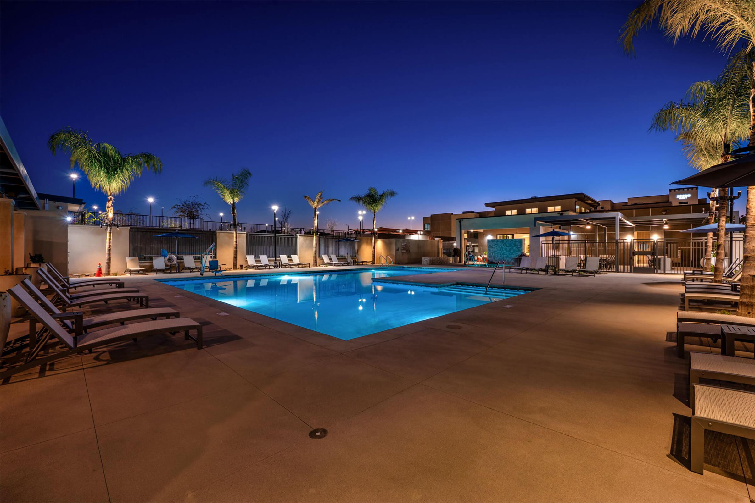 A well-lit outdoor swimming pool surrounded by lounge chairs, palm trees, and softly glowing lights at dusk. The pool area features a modern design, with comfortable seating and a serene atmosphere, ideal for relaxation and evening gatherings.