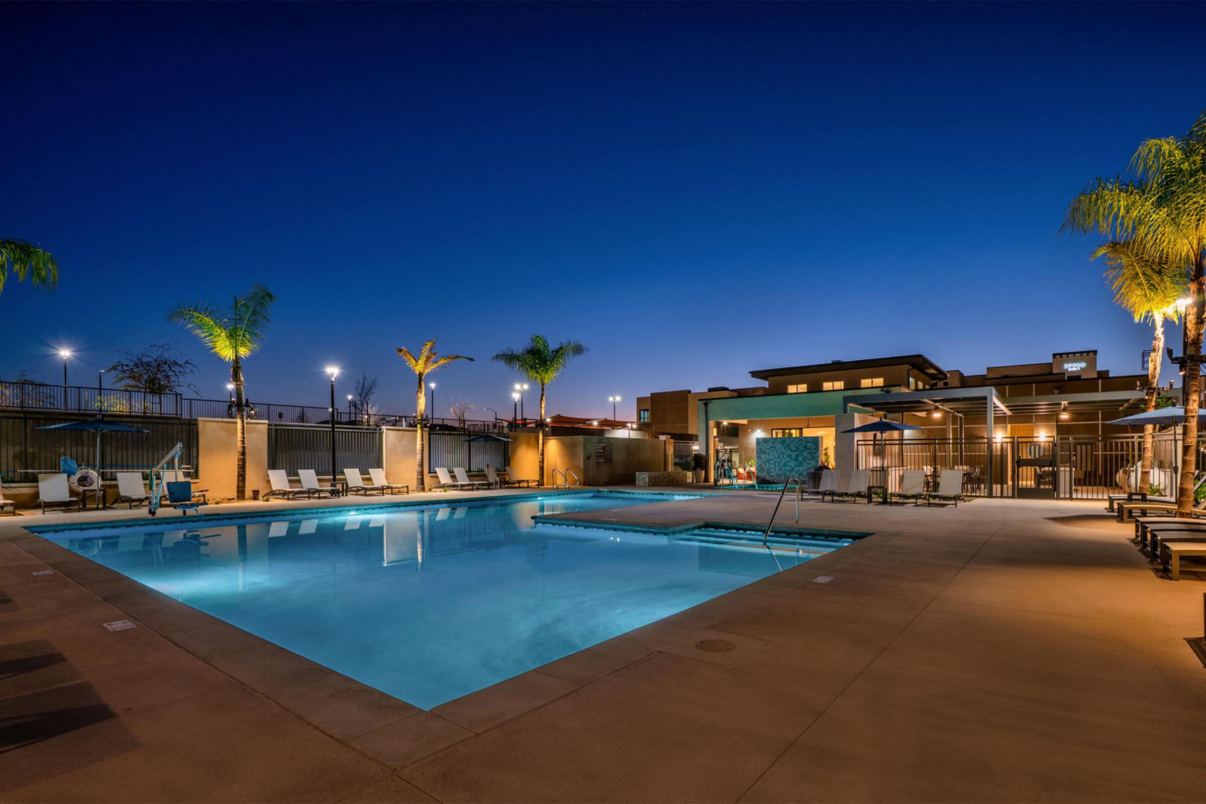 A spacious outdoor swimming pool illuminated at night, surrounded by palm trees and lounge chairs. The pool features a shallow area and is set against a backdrop of modern buildings. Soft lighting enhances the tranquil atmosphere, creating a serene and inviting space for relaxation.