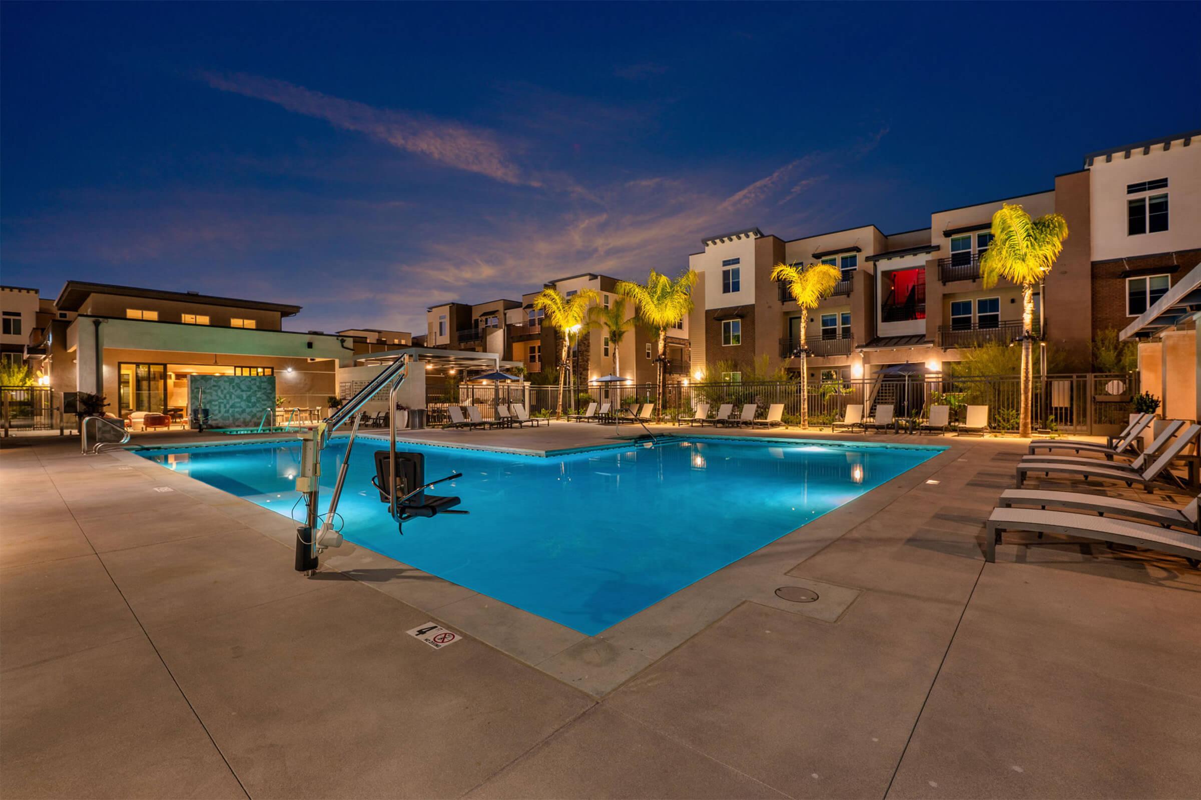 A beautifully lit swimming pool area at dusk, surrounded by modern apartment buildings. Palm trees add a tropical touch, and lounge chairs are lined up beside the pool. The atmosphere is calm and inviting, perfect for relaxation.