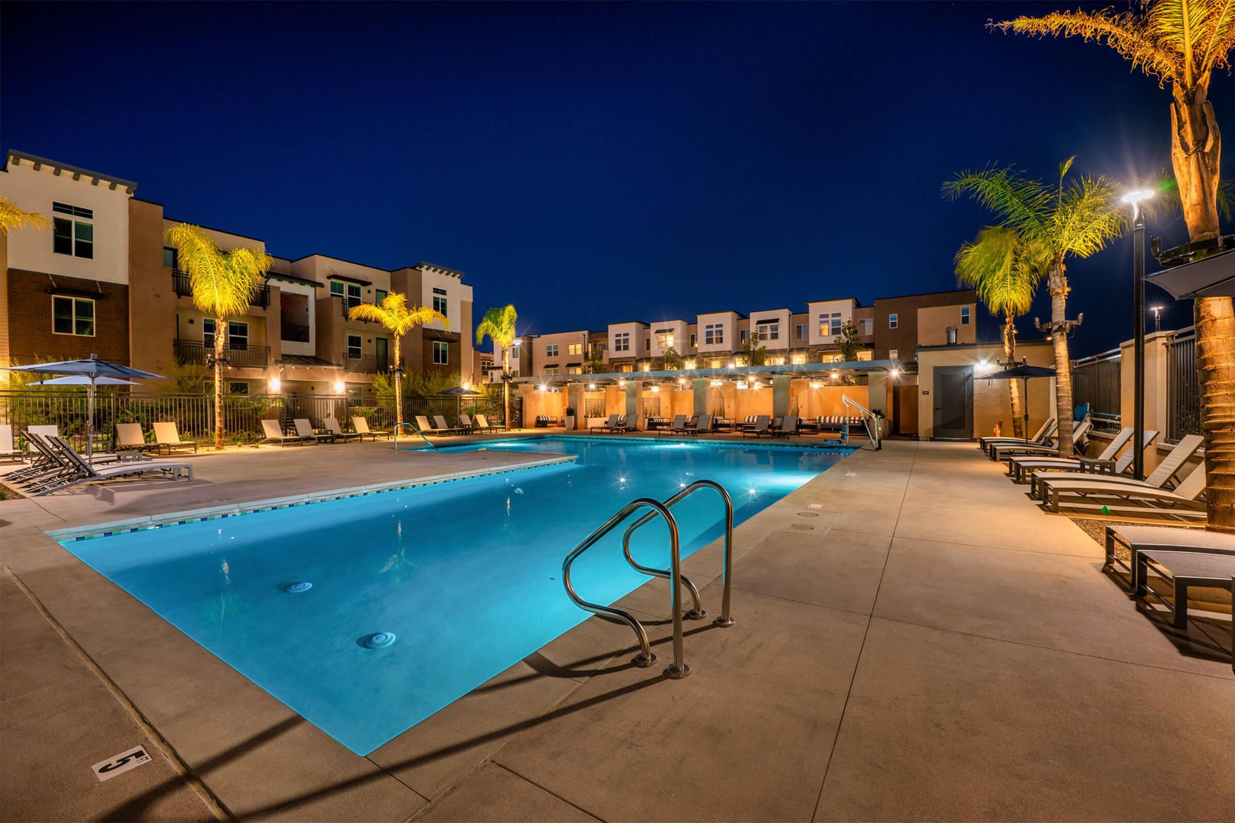 A well-lit swimming pool at night surrounded by lounge chairs and palm trees. The pool area features elegant lighting and modern apartment buildings in the background, creating a relaxing and inviting atmosphere.
