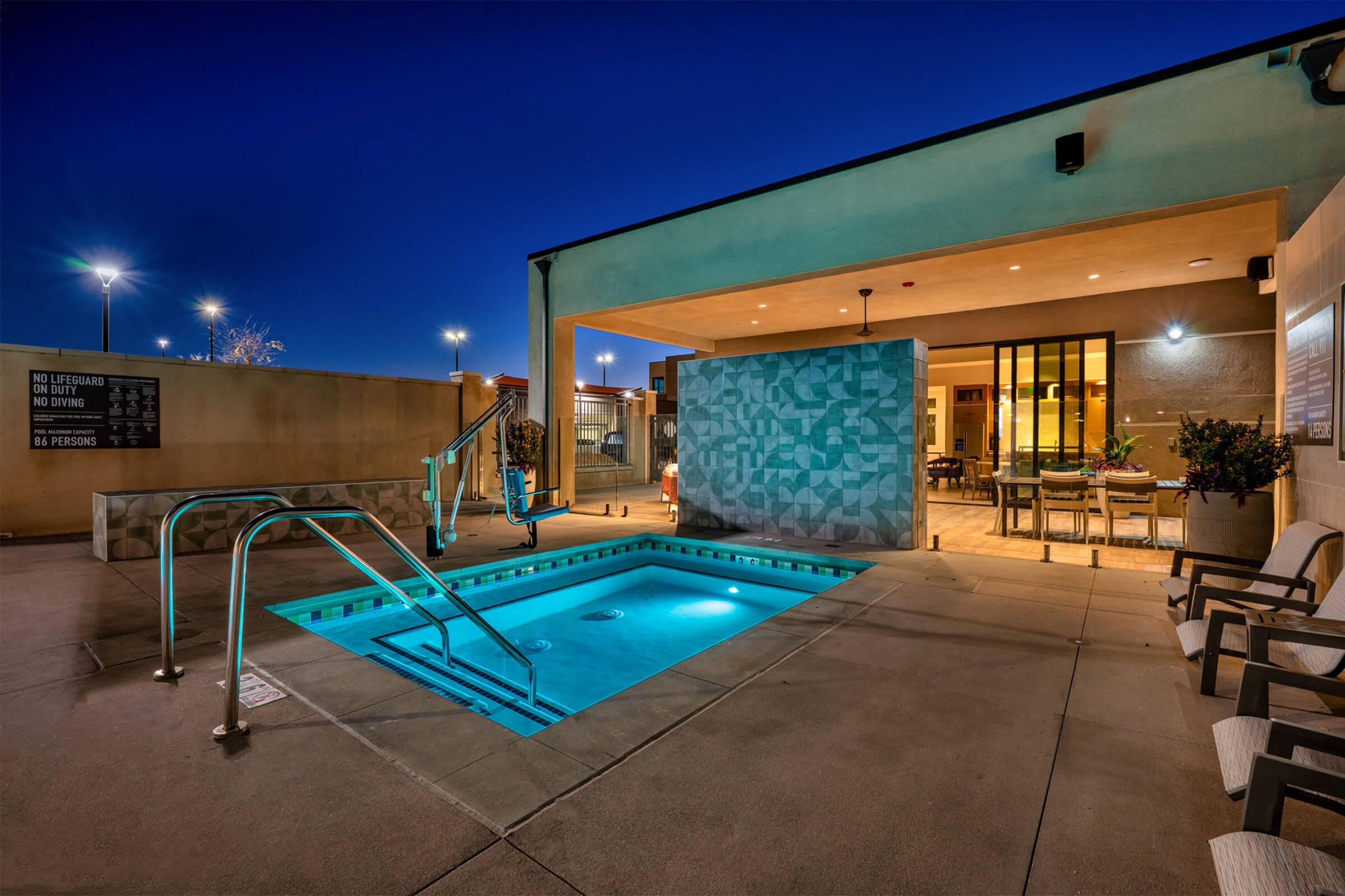 A well-lit outdoor pool area at night, featuring a small hot tub, sleek lounge chairs, and a shaded seating space with a modern design. Soft lighting enhances the ambiance, while nearby signs provide safety information. The surrounding area includes a concrete surface and decorative wall.